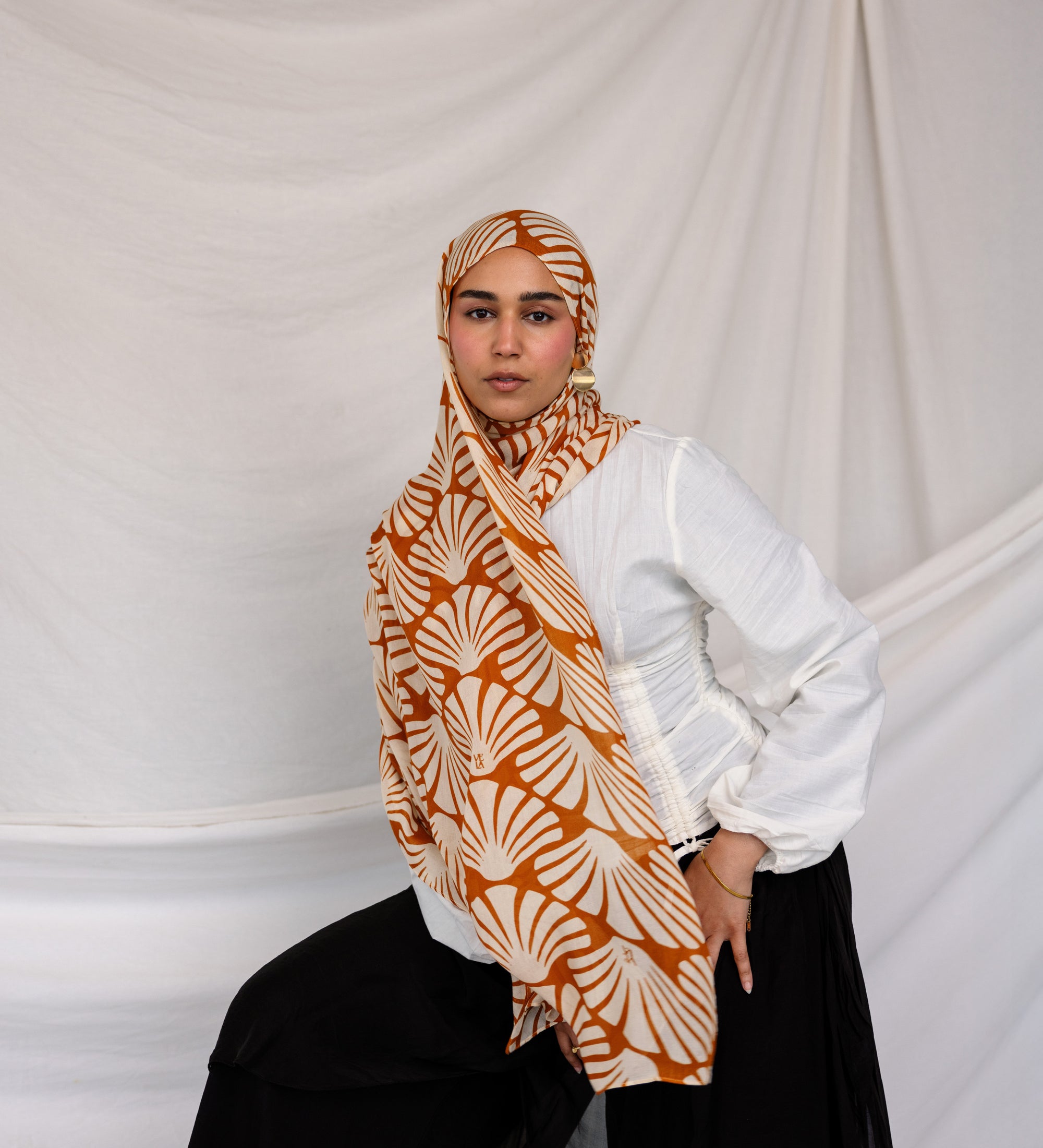 Woman wearing a Amber Shells printed hijab, white shirt, and black pants against a plain background.
