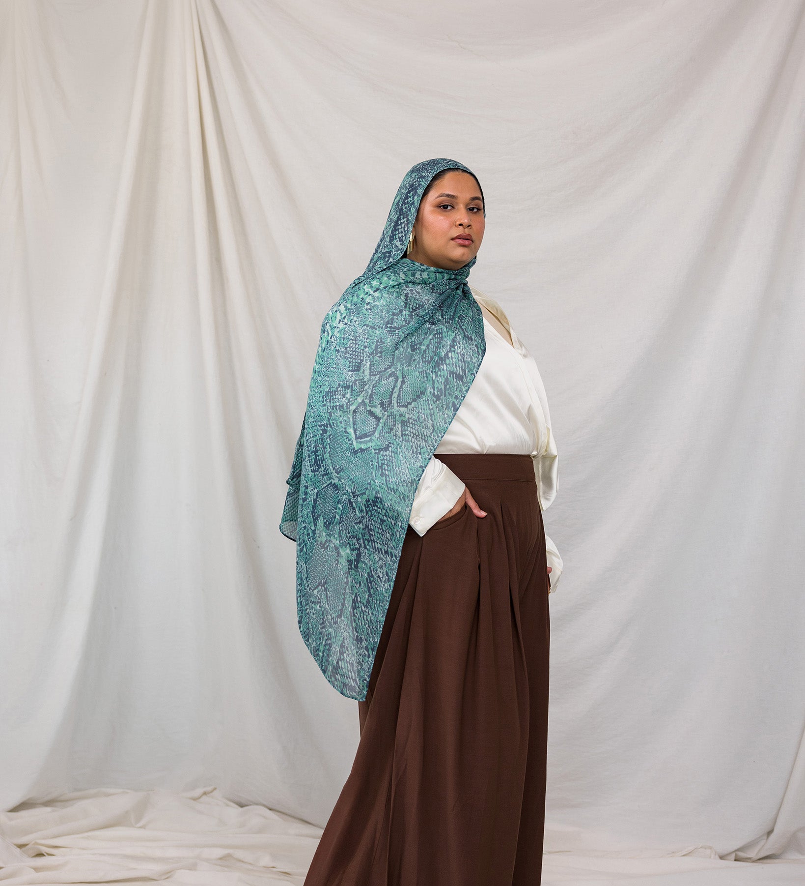 A model posing in the Aqua Cobra scarf, a teal and blue snakeskin print scarf. Model is also wearing the VELA Palazzo pant.