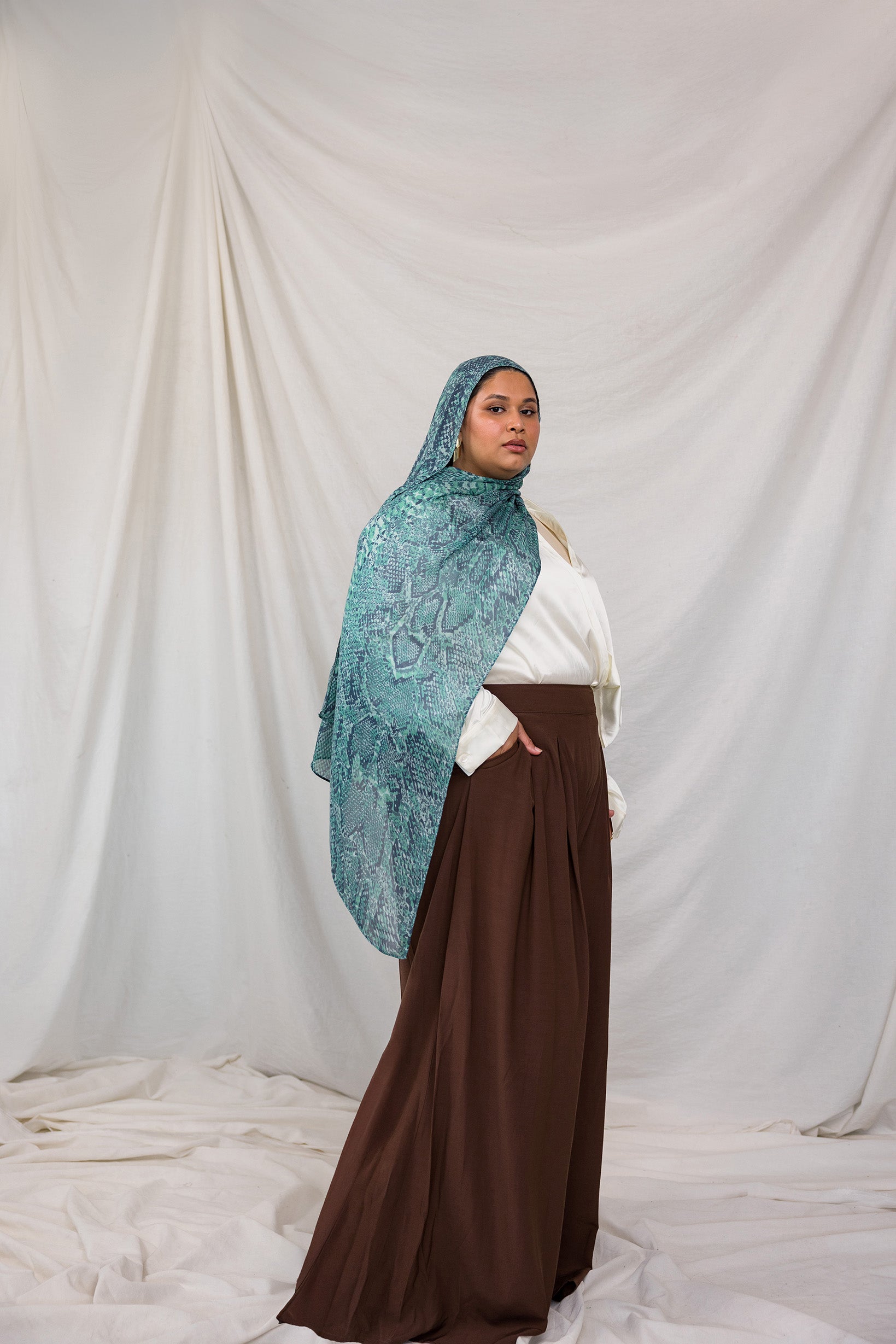 A model posing in the Aqua Cobra scarf, a teal and blue snakeskin print scarf. Model is also wearing the VELA Palazzo pant.