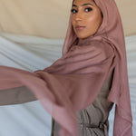 A woman wearing a blush pink modal hijab with a slight purple undertone, draping elegantly over her head and shoulders.