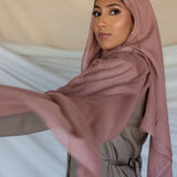 A woman wearing a blush pink modal hijab with a slight purple undertone, draping elegantly over her head and shoulders.