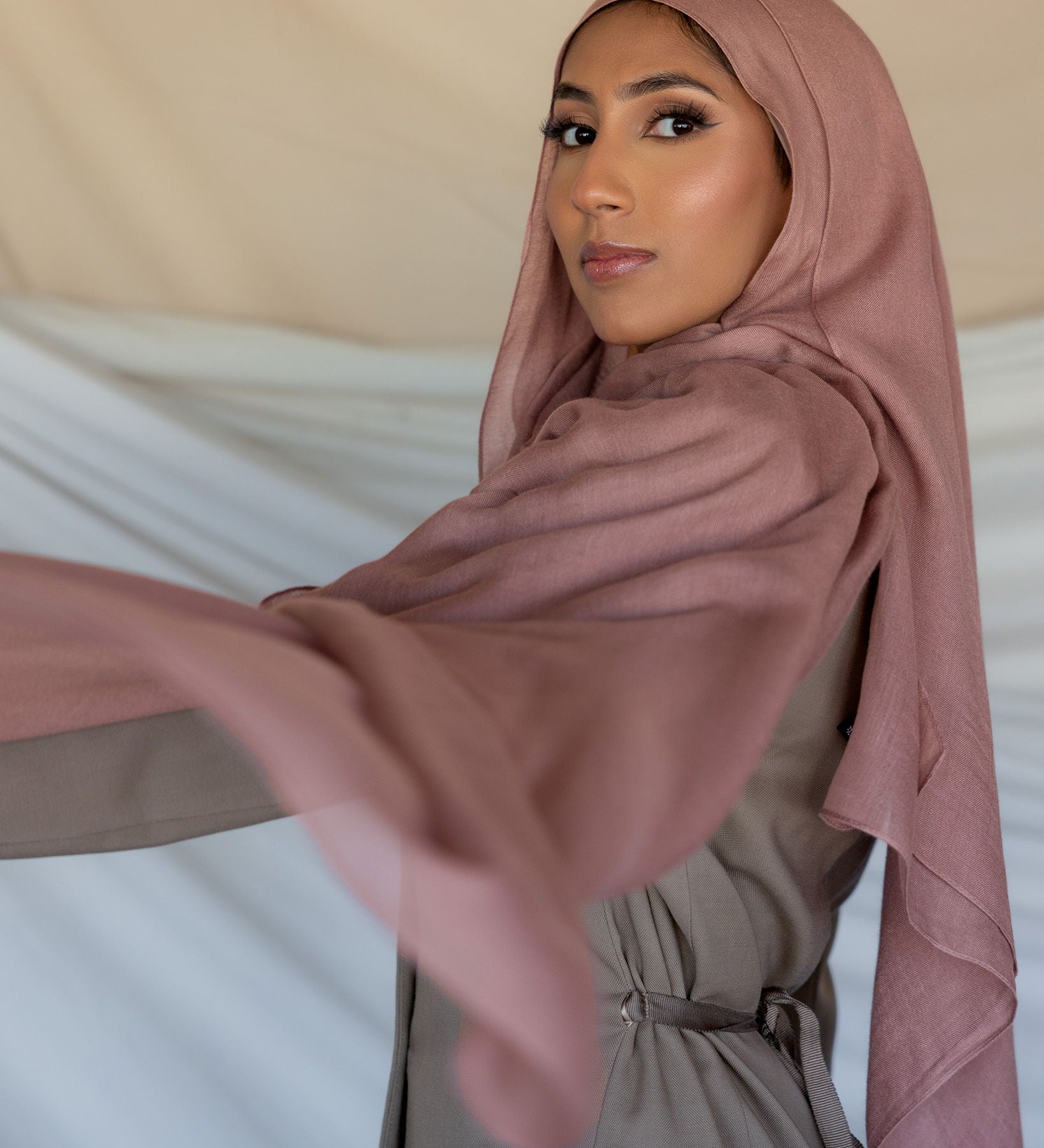 A woman wearing a blush pink modal hijab with a slight purple undertone, draping elegantly over her head and shoulders.