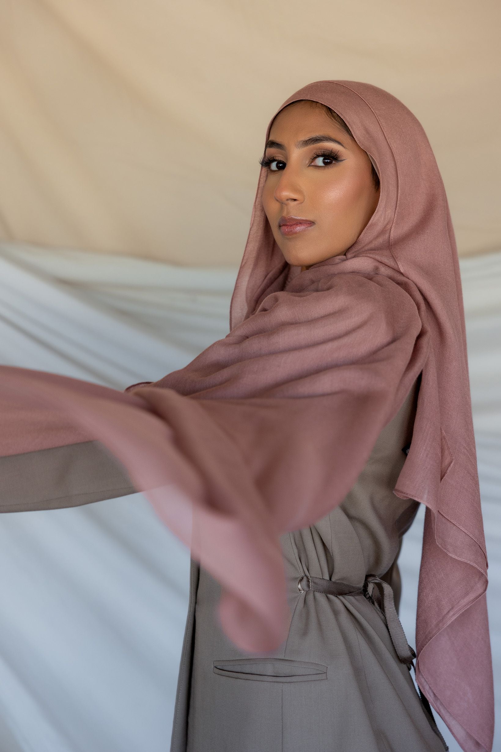 A woman wearing a blush pink modal hijab with a slight purple undertone, draping elegantly over her head and shoulders.