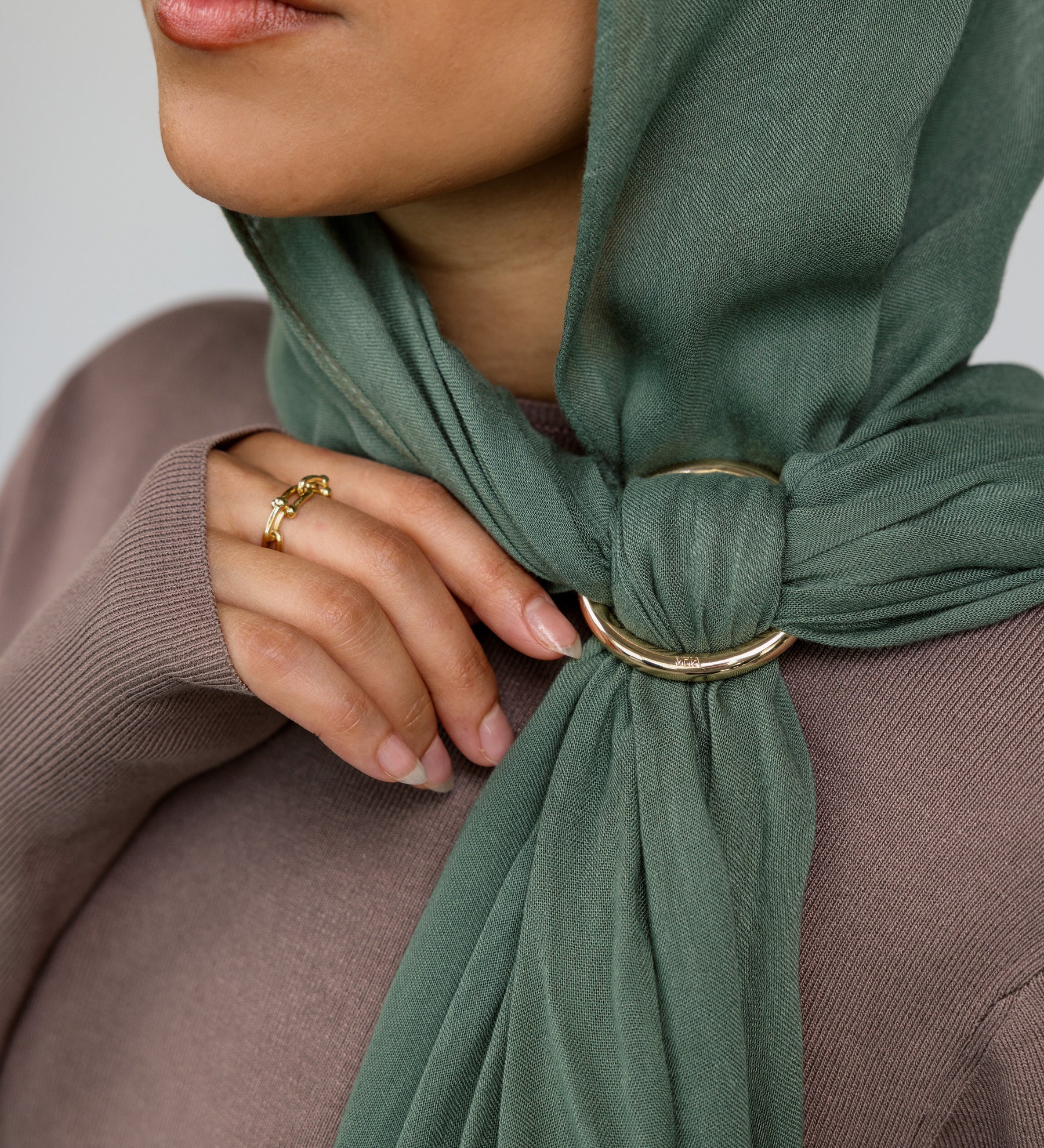 Person wearing a green hijab with a gold hijab hoop, against a neutral background.