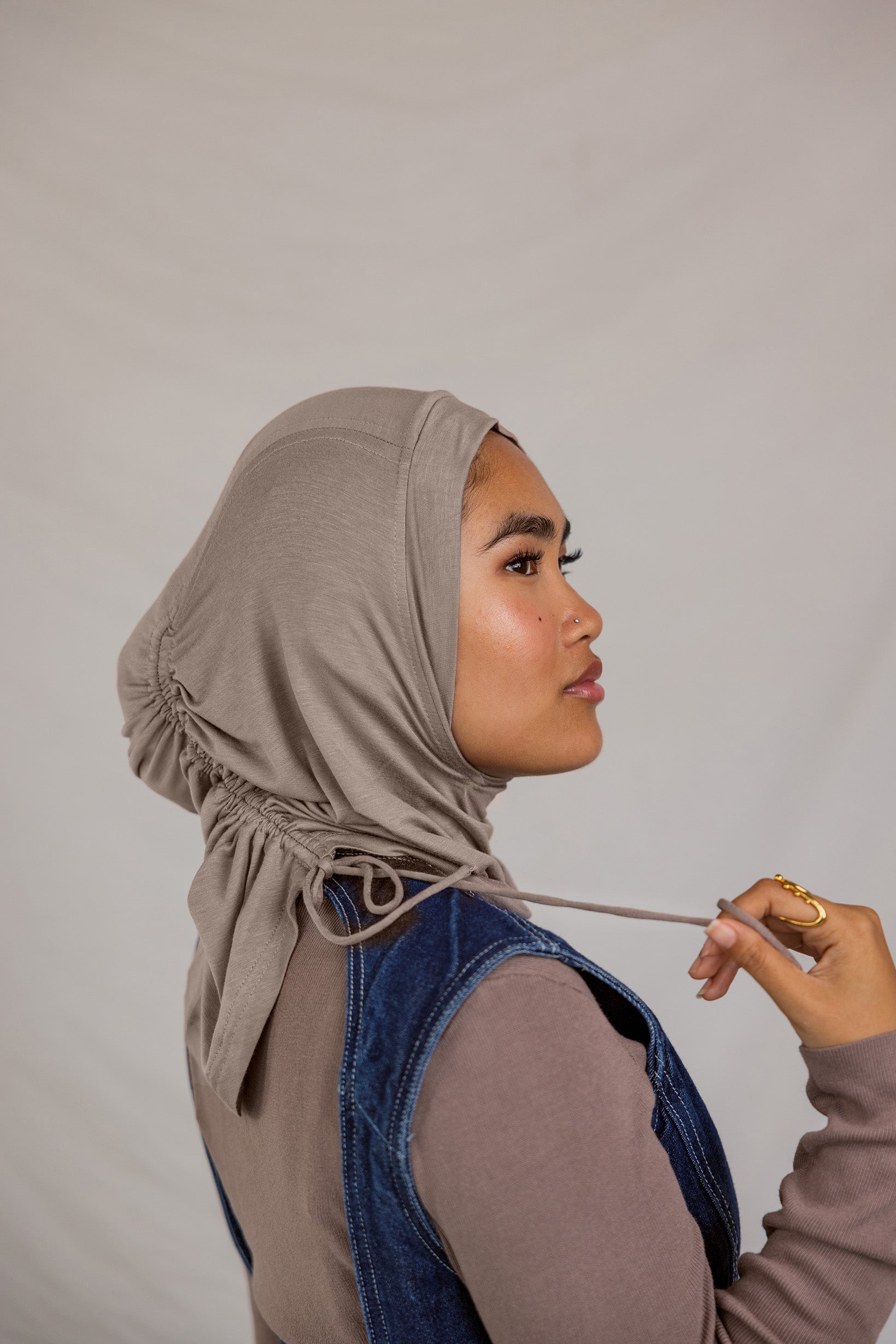 Woman adjusting a taupe grey ruching hijab against a plain background.