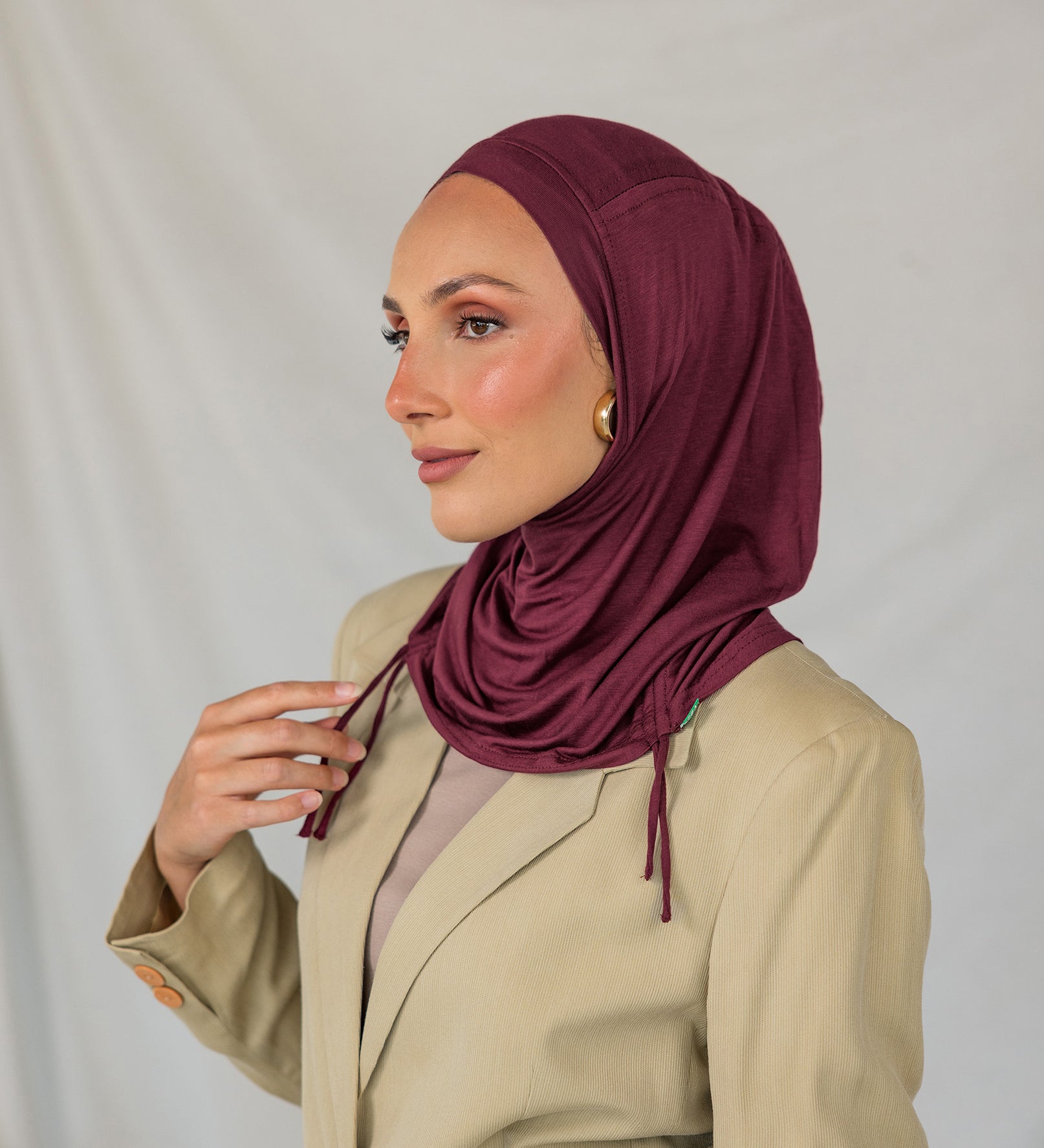 Woman wearing a burgundy ruched hijab and beige blazer against a plain background.