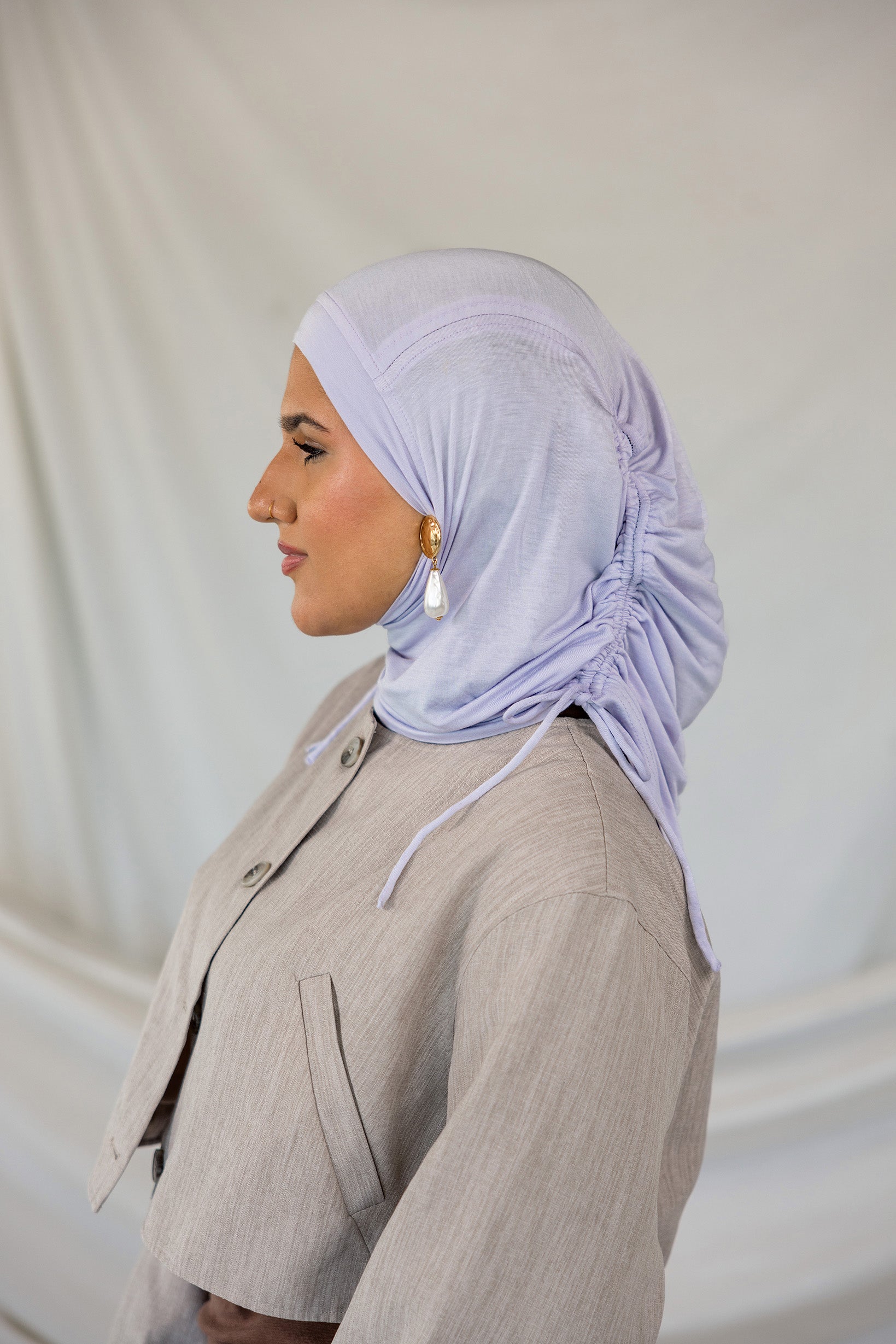 Woman wearing a lavender ruching hijab and beige suit against a plain background.