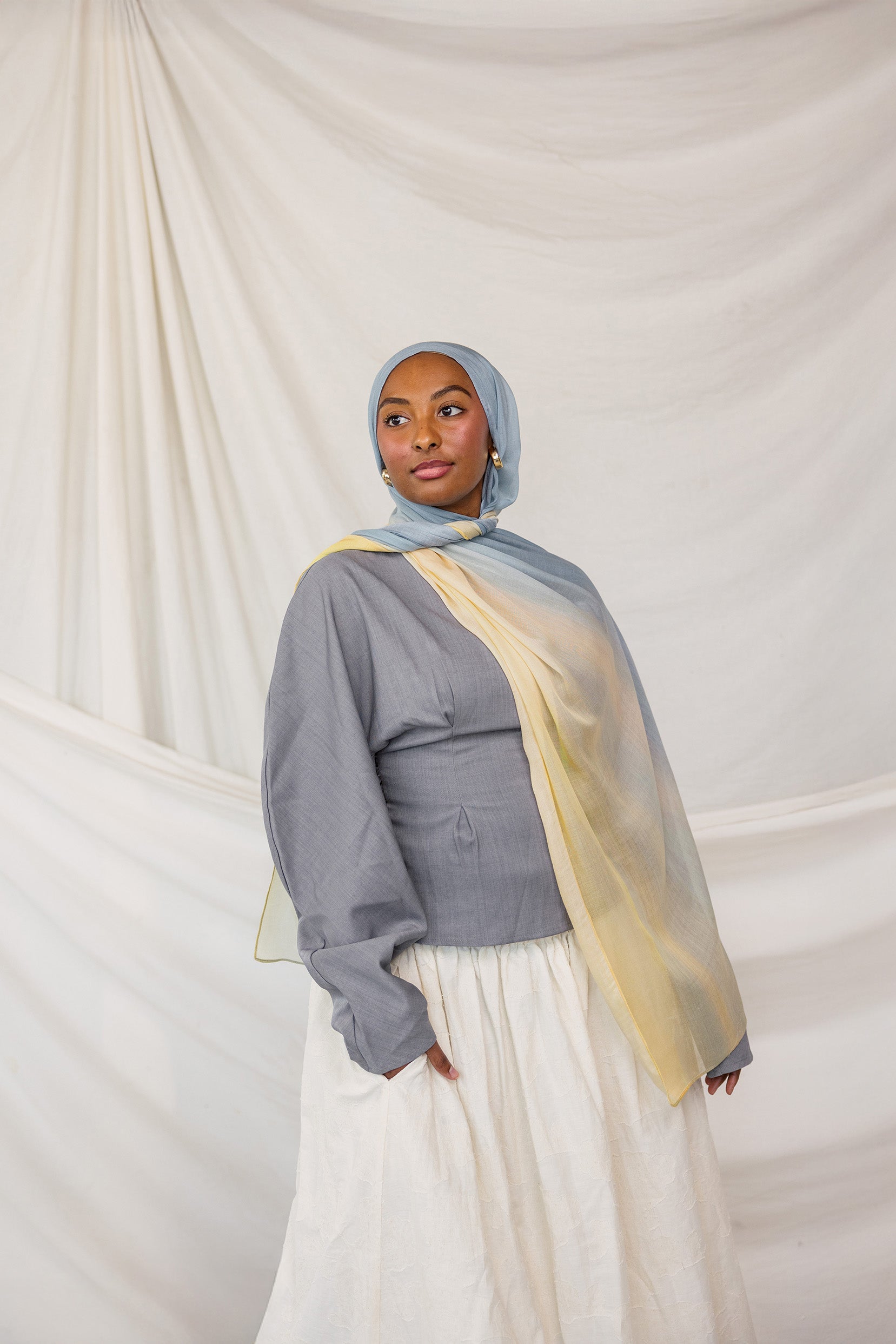 Model wearing a gray top, white skirt, and blue to yellow gradient hijab against a plain background