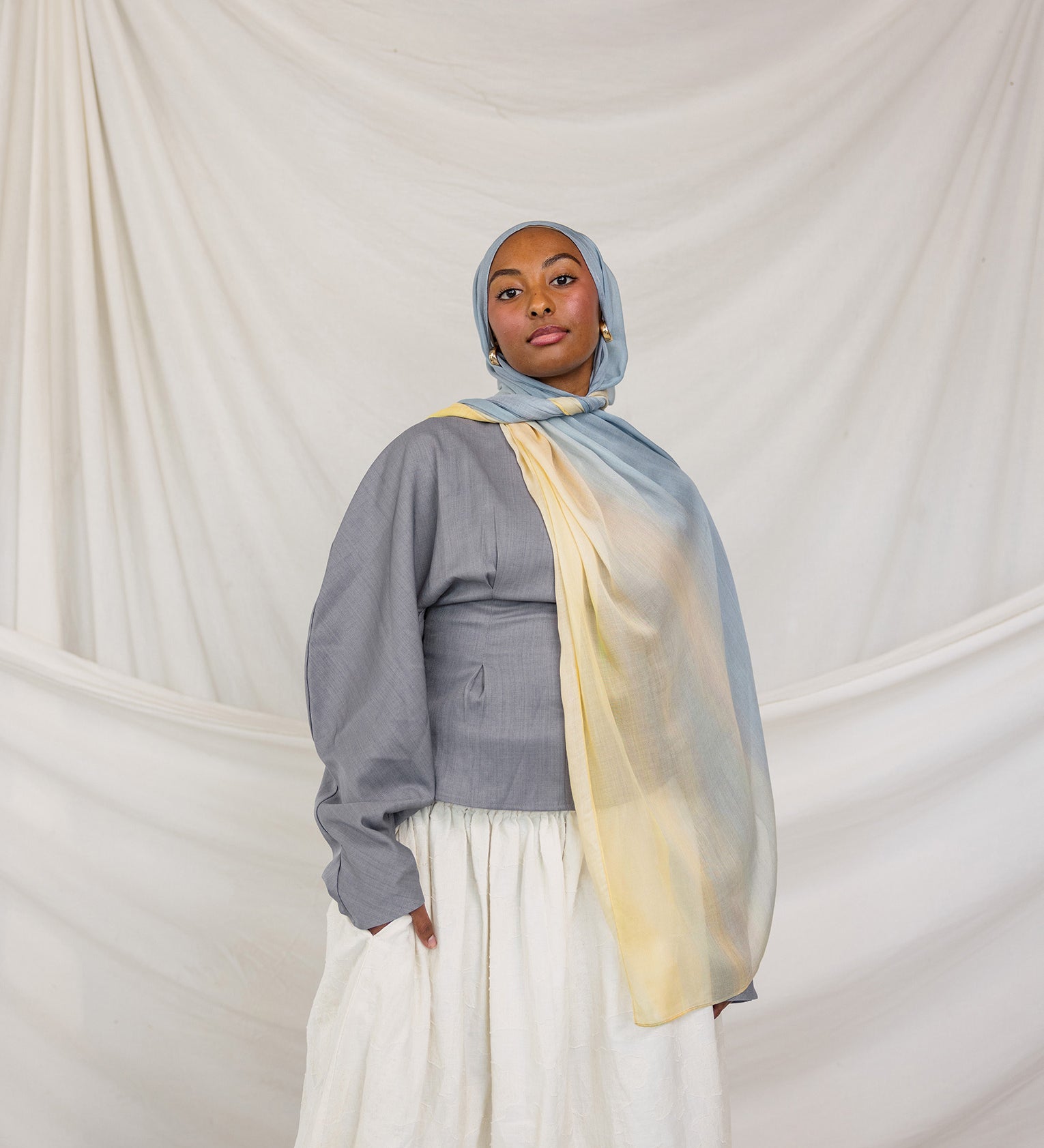Person wearing a gray top, white skirt, and colorful blue / yellow hijab against a plain background