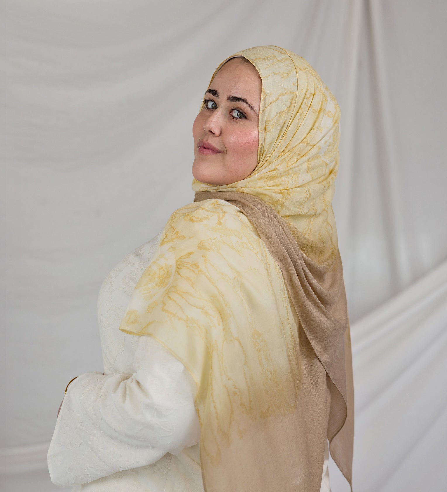 Woman wearing a yellow beige shell printed hijab and white outfit against a plain background