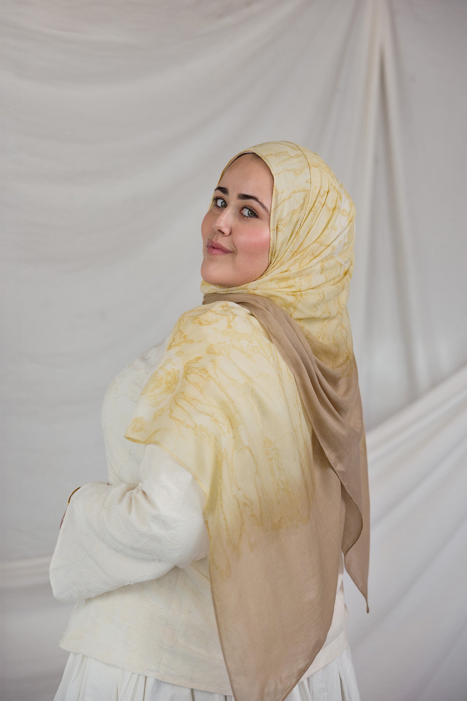 Woman wearing a yellow beige shell printed hijab and white outfit against a plain background