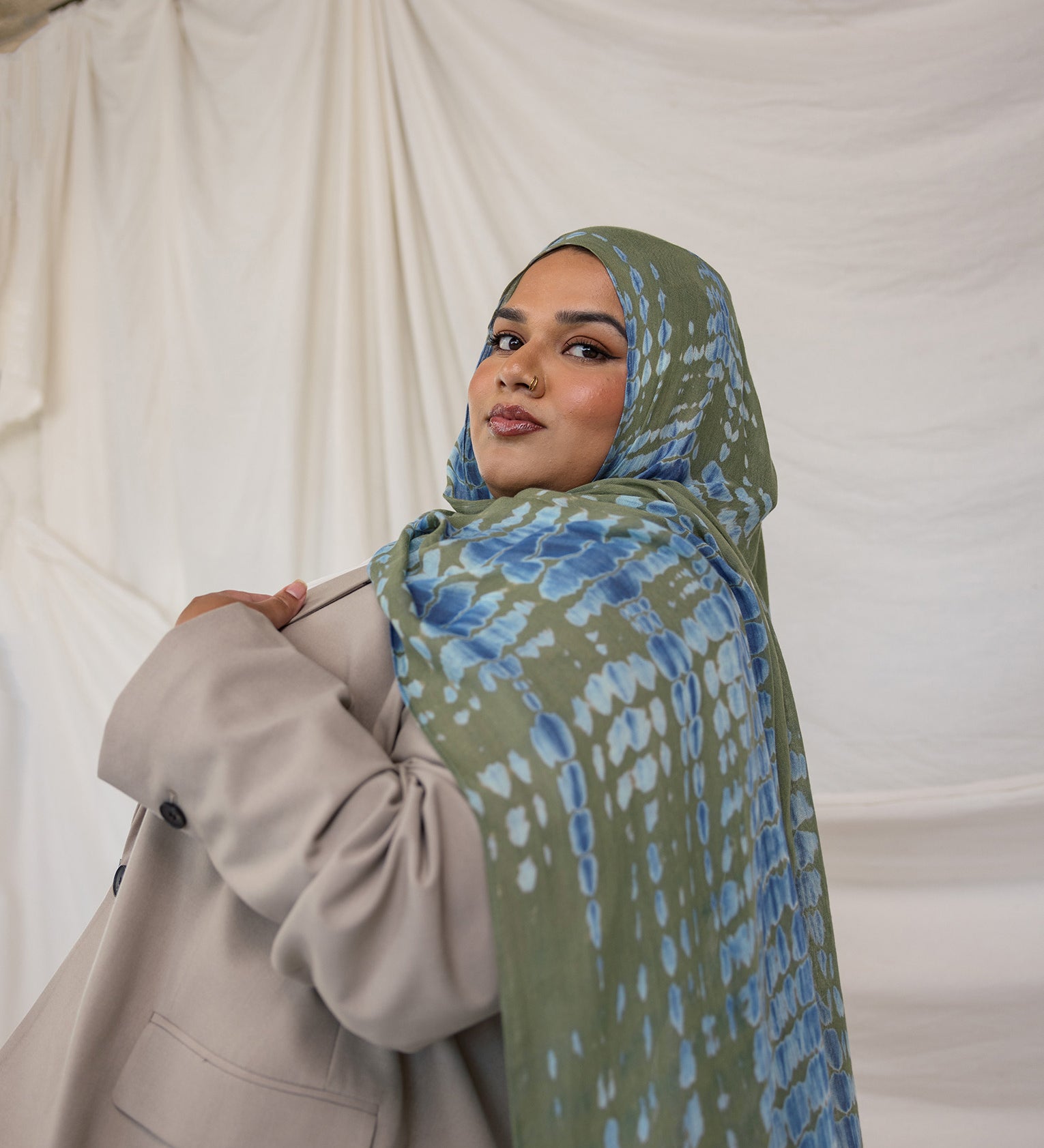 Woman wearing a green and blue ripple patterned hijab and beige coat against a white curtain background