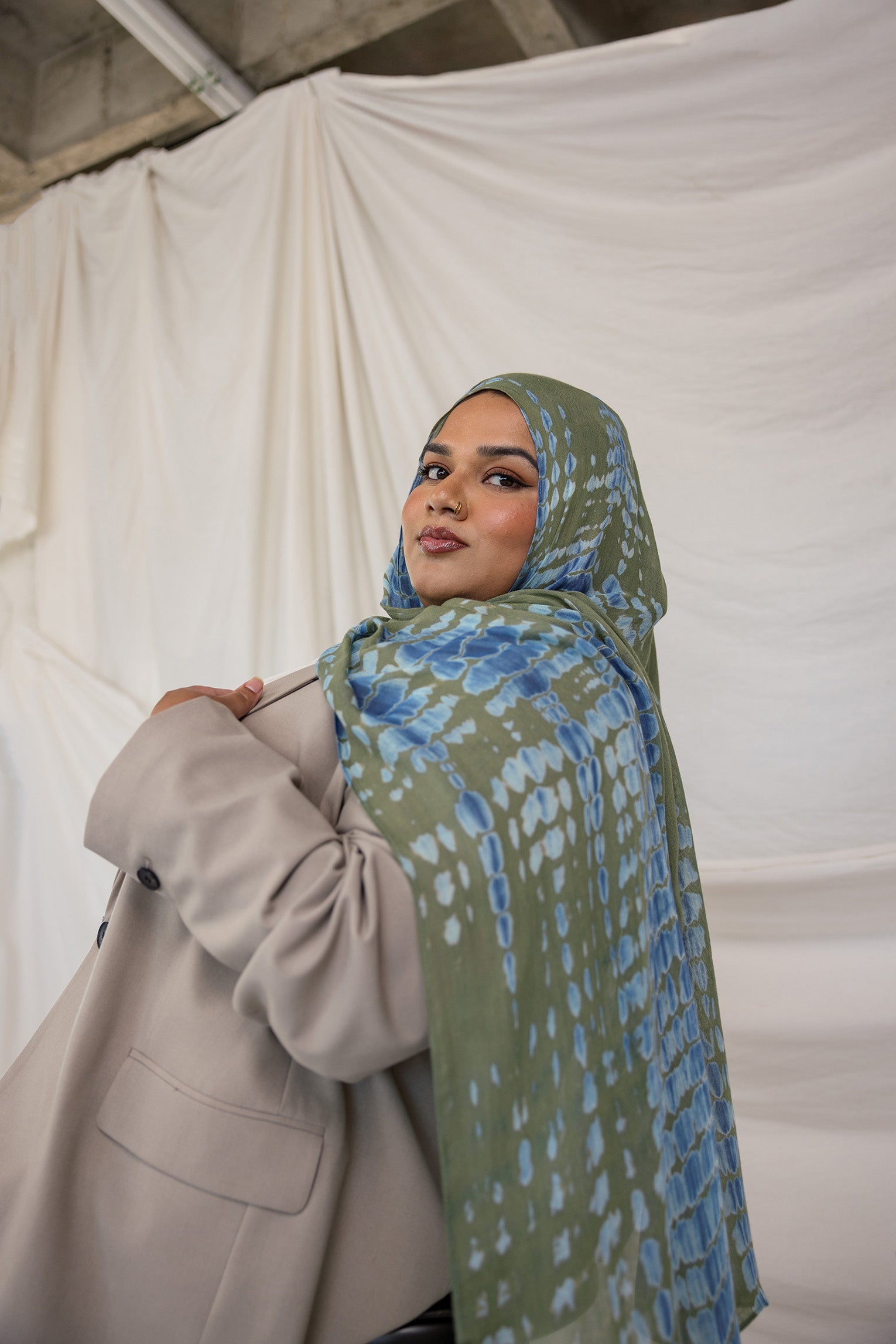 Woman wearing a green and blue ripple patterned hijab and beige coat against a white curtain background