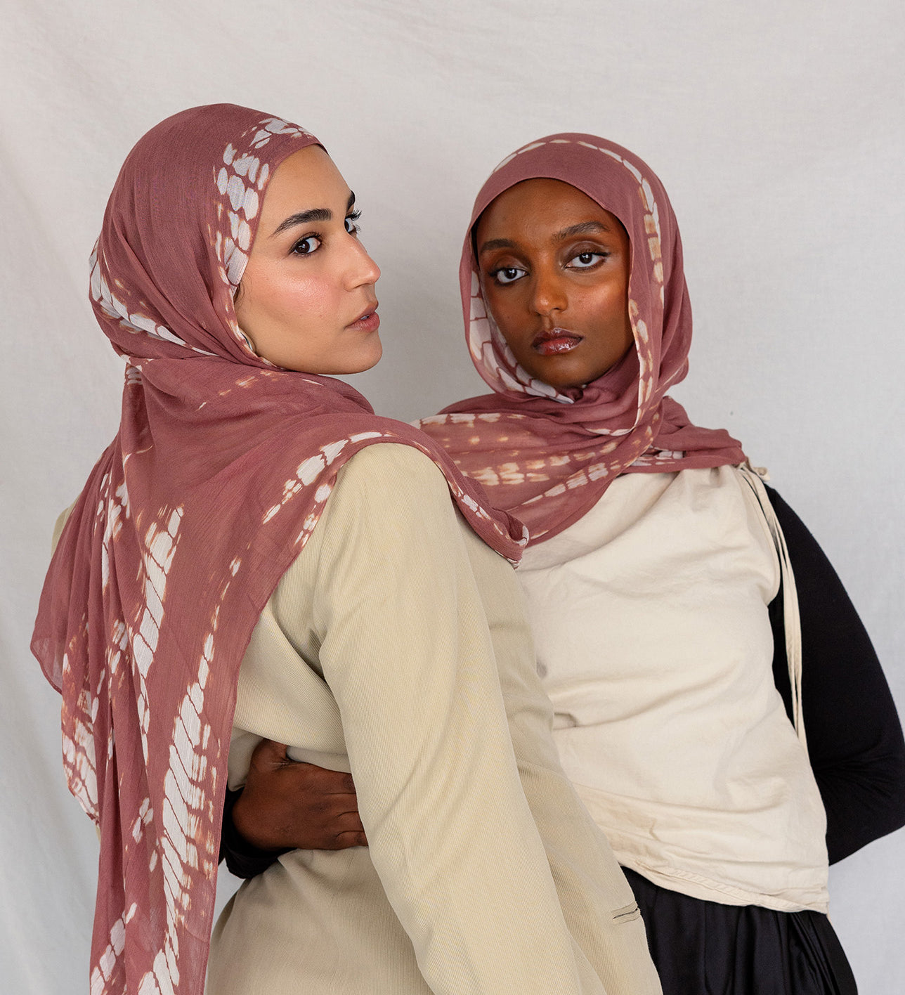 Two women wearing pink rippled printed hijabs and neutral-colored clothing against a plain background