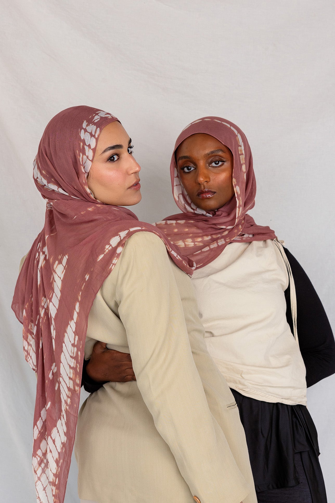 Two women wearing pink rippled printed hijabs and neutral-colored clothing against a plain background