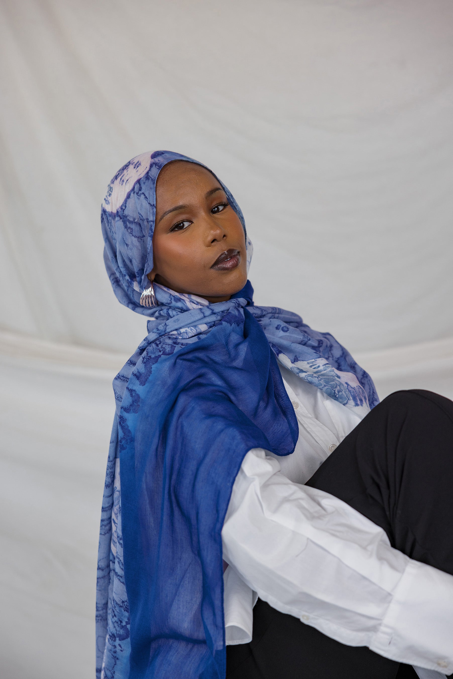 Model wearing a blue hijab with white shell prints and sitting on a white surface