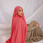 Woman wearing a bright coral hijab and beige outfit sitting on a white fabric background