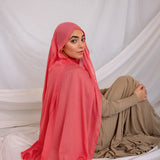 Woman wearing a bright coral hijab and beige outfit sitting on a white fabric background