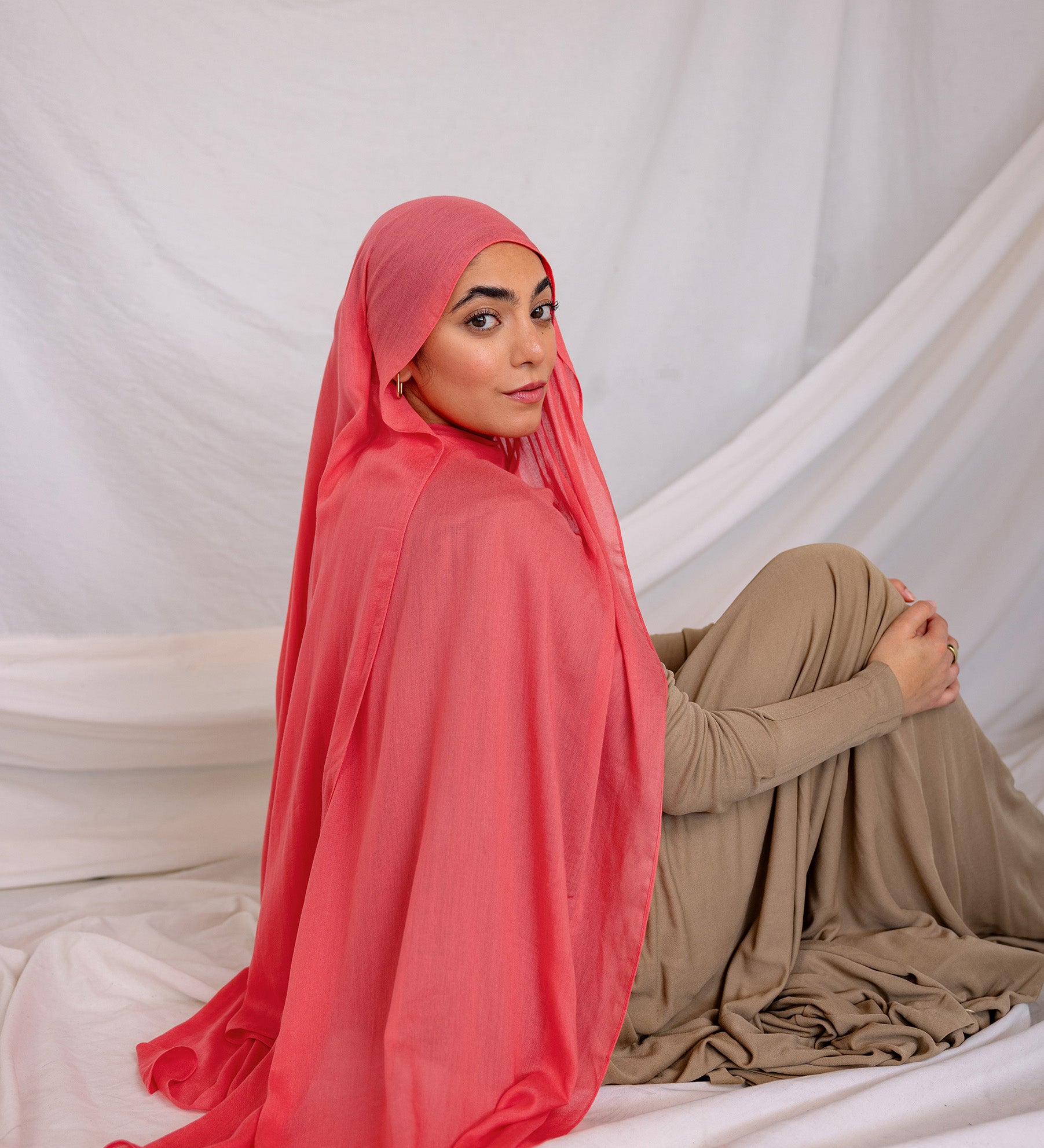 Woman wearing a bright coral hijab and beige outfit sitting on a white fabric background