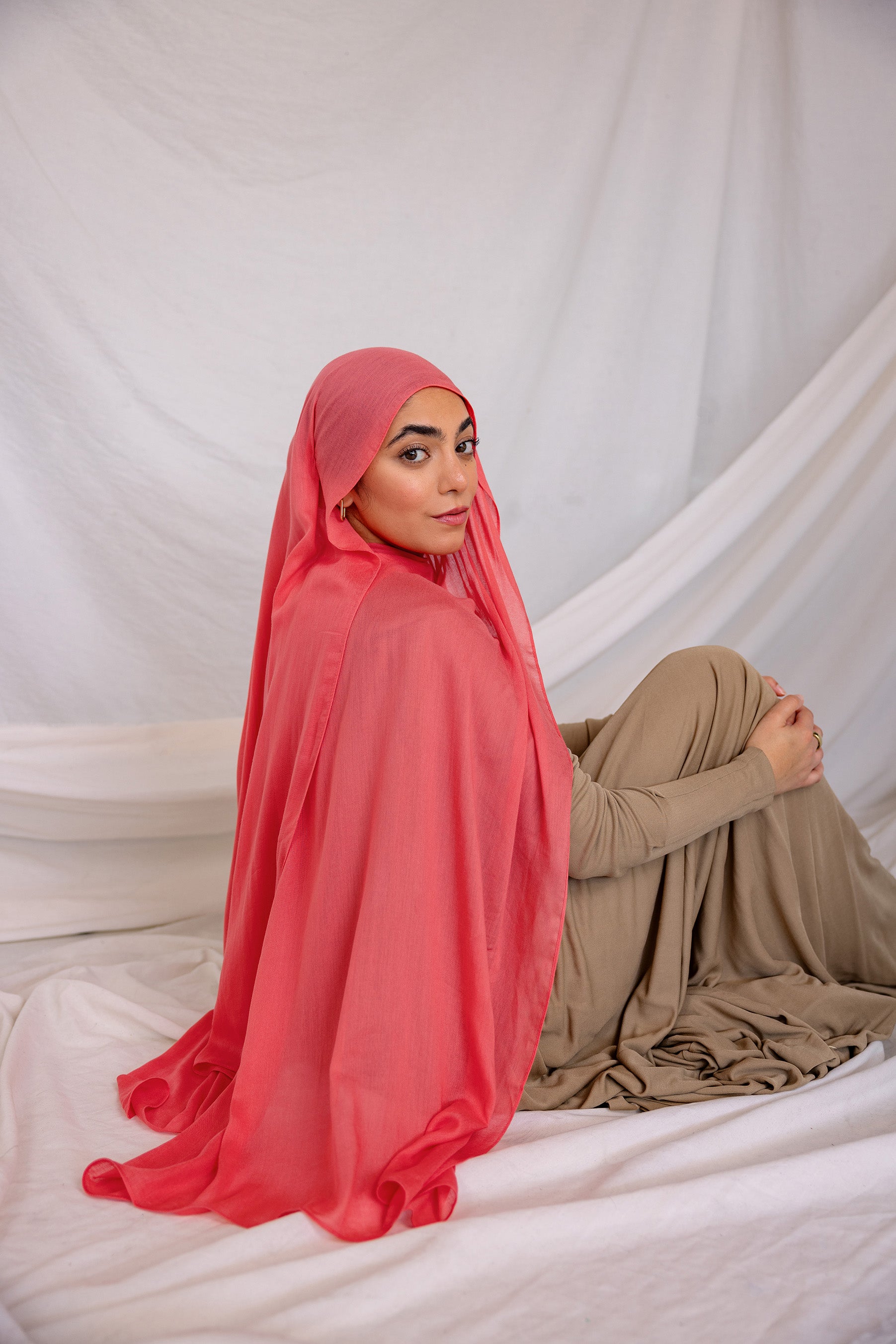 Woman wearing a bright coral hijab and beige outfit sitting on a white fabric background
