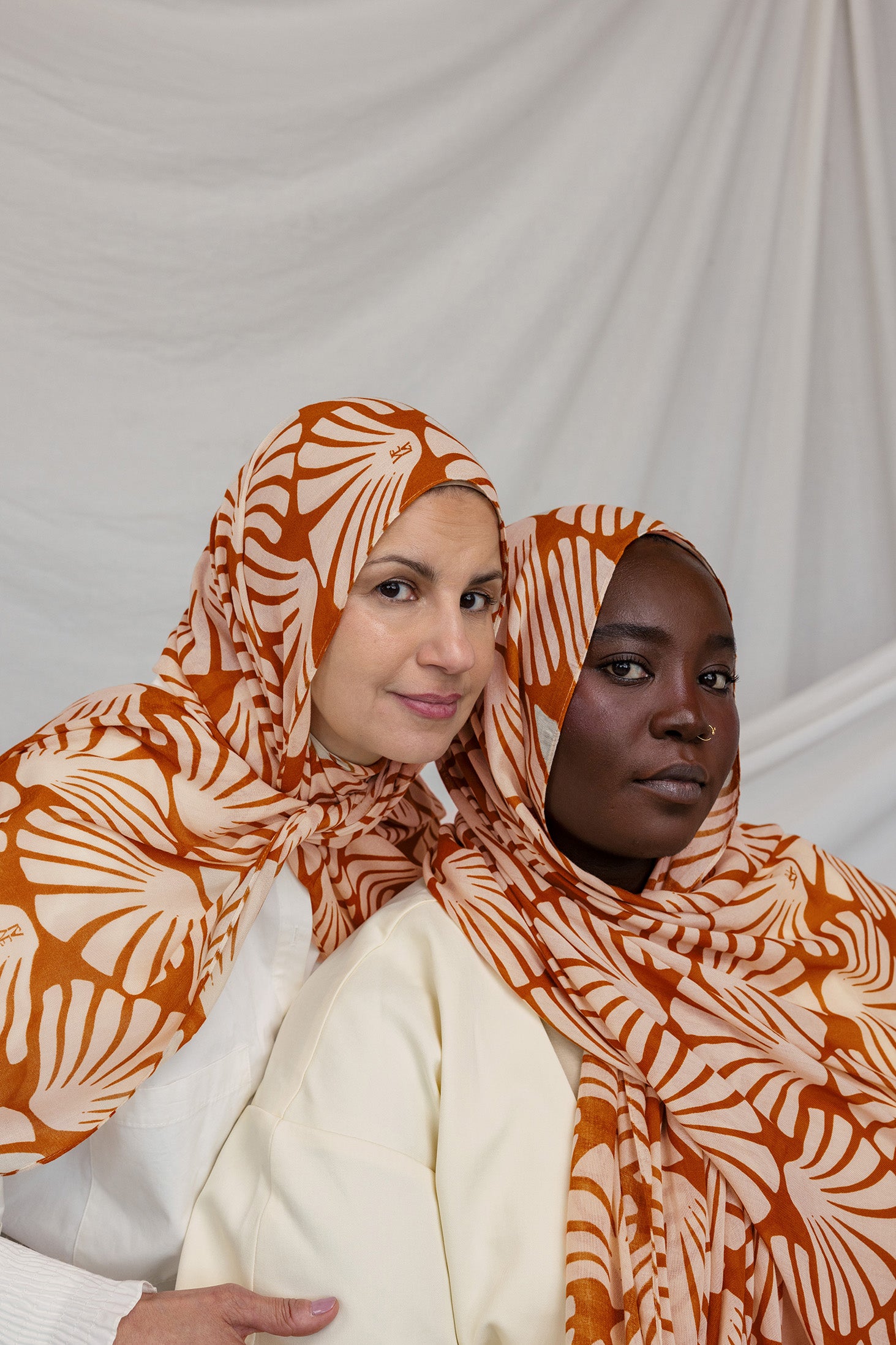 Two women wearing orange and white Amber Shells patterned hijabs against a plain background