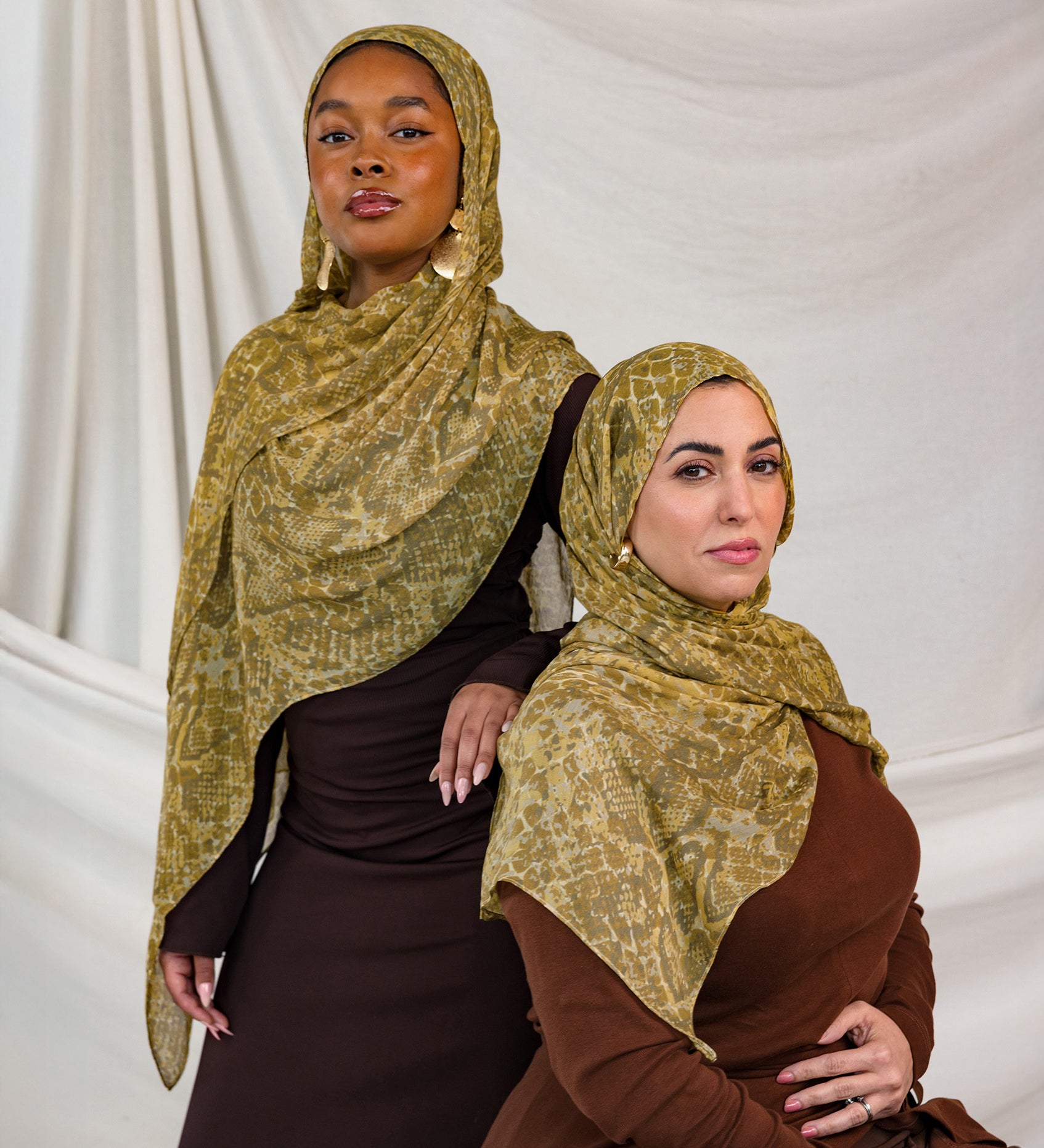 Models shown posing in the Golden Cobra scarf by VELA. This image features one model standing and one sitting, both wearing the golden/yellow toned snakeskin print.