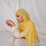 Woman wearing a bright yellow hijab and white outfit sitting on a white draped background