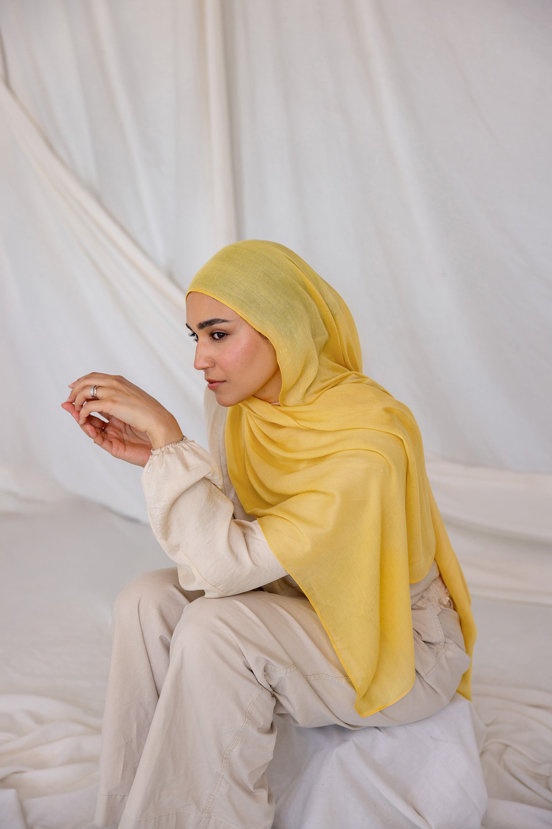 Woman wearing a bright yellow hijab and white outfit sitting on a white draped background