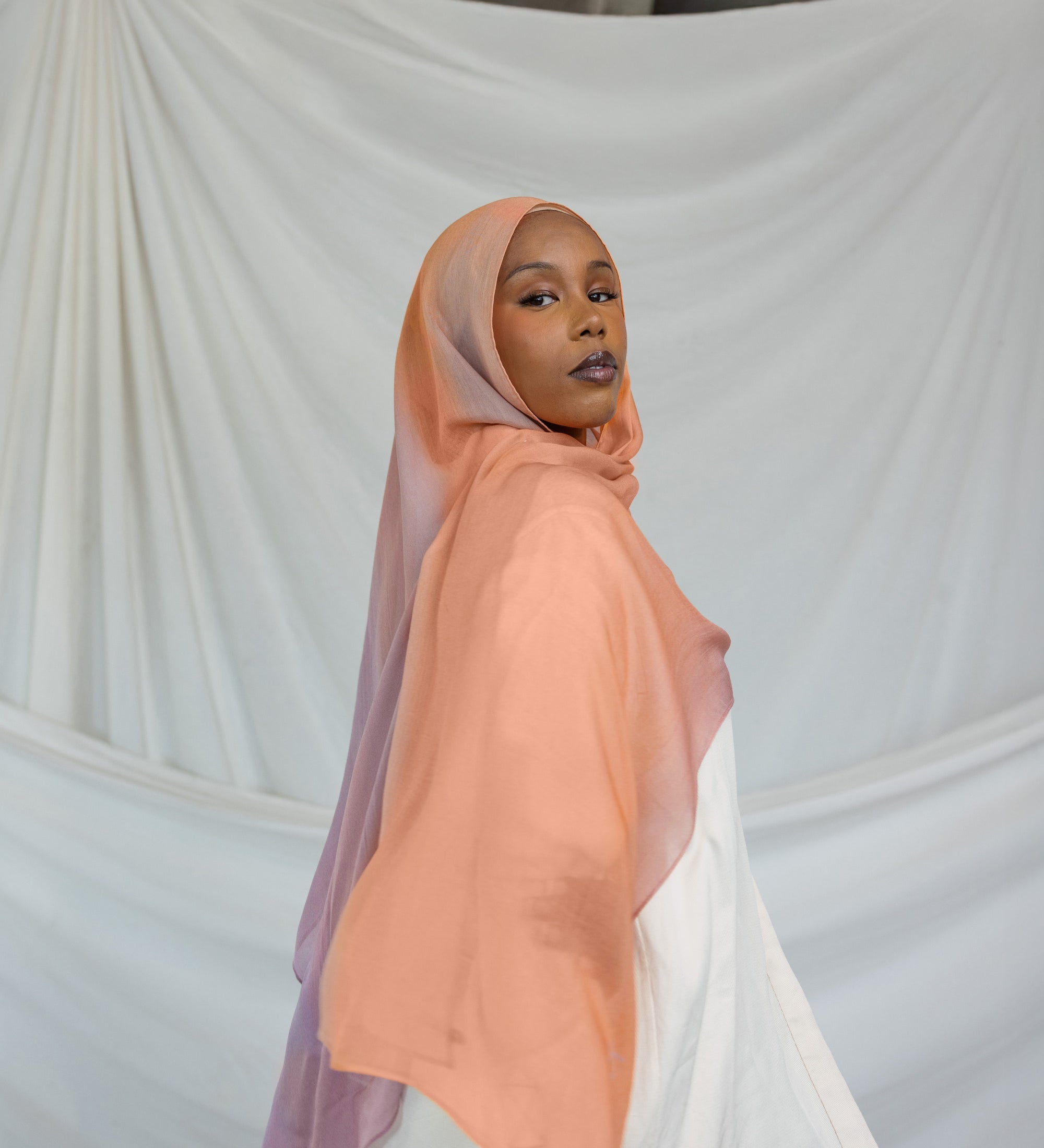 Woman wearing a bright pink / purple pastel hijab in front of draped fabric
