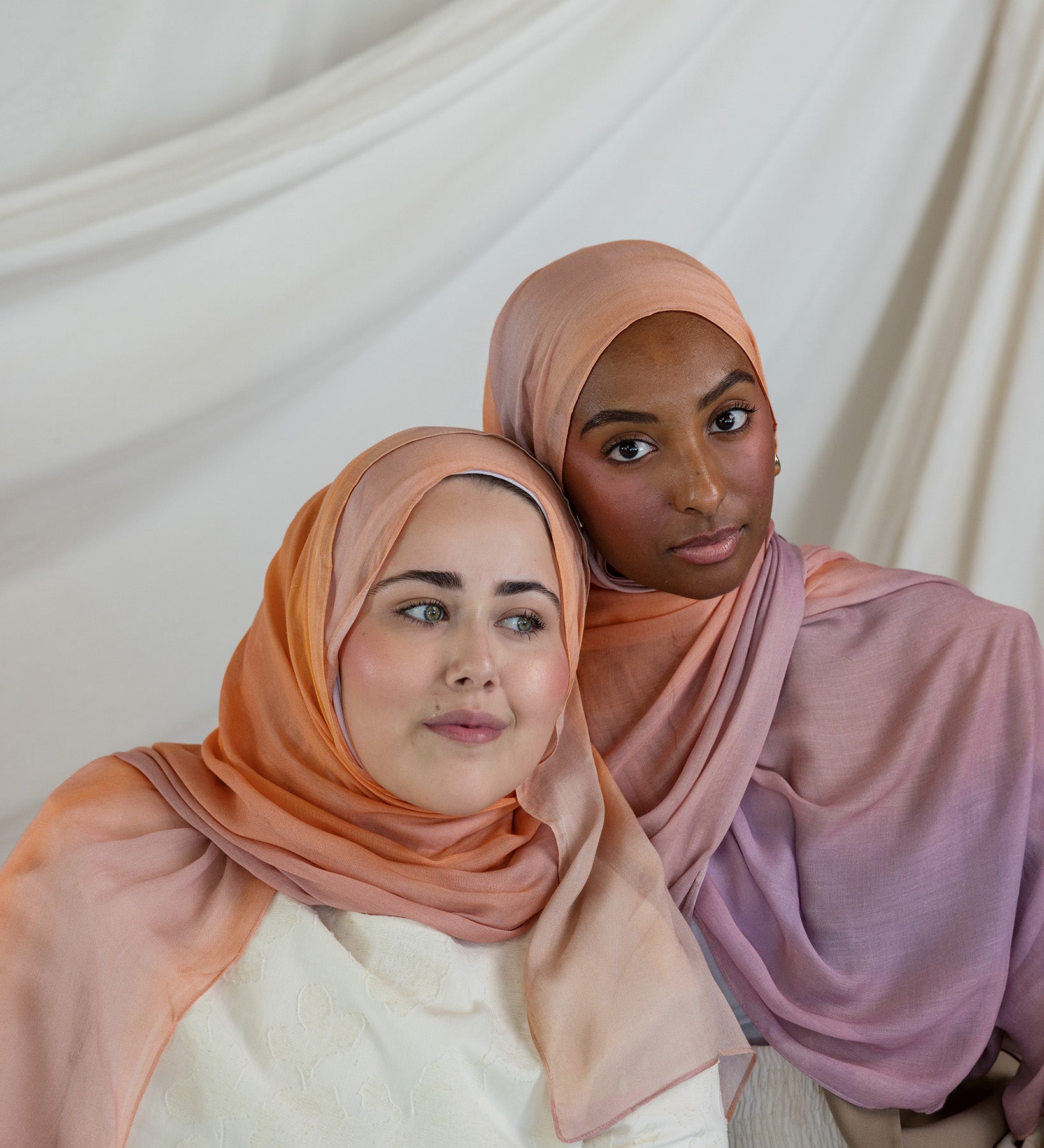 Two women wearing pink and purple ombre hijabs against a white curtain background