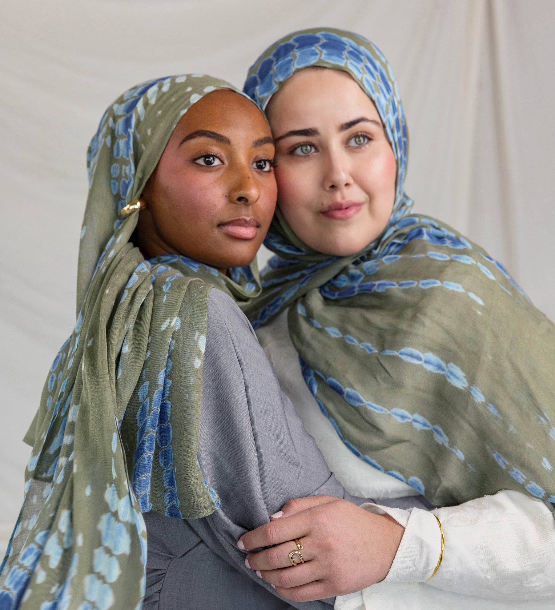 Two women wearing green and blue patterned headscarves embracing each other against a plain background