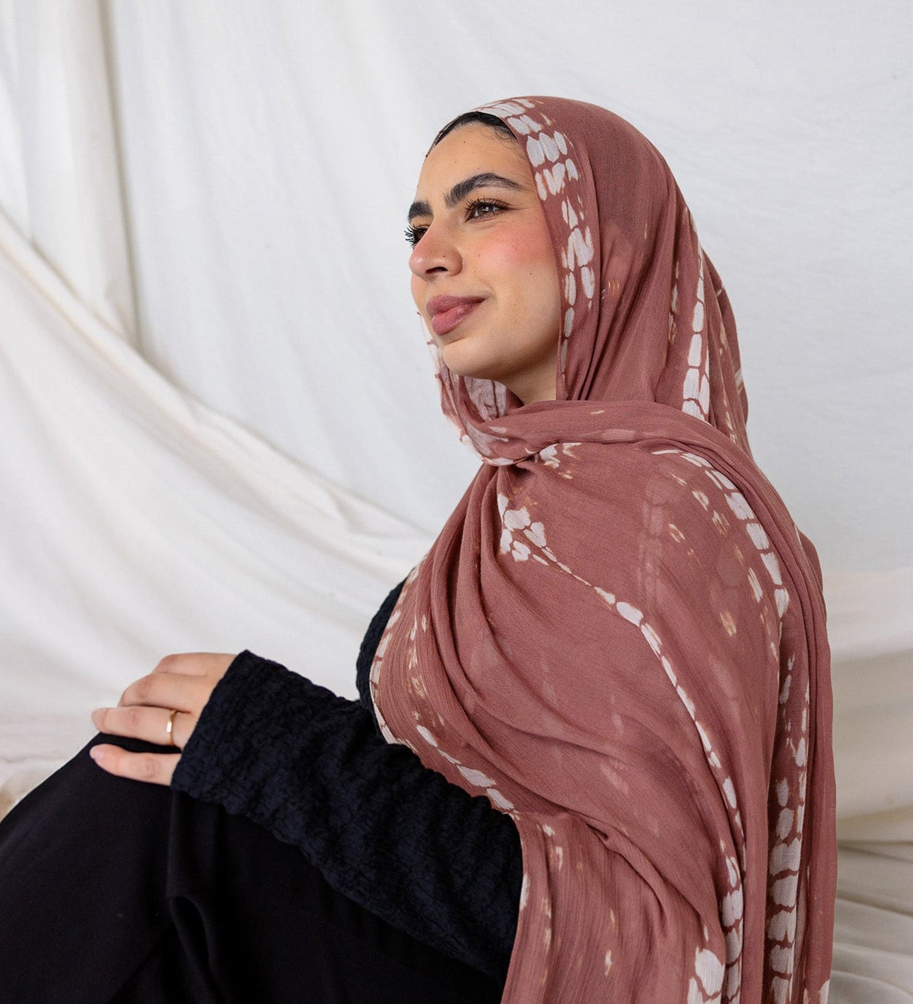 Woman wearing a patterned ripple pink printed hijab sitting against a white curtain.
