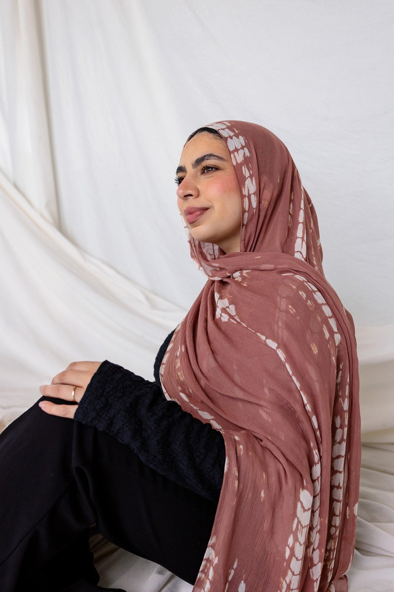 Woman wearing a patterned ripple pink printed hijab sitting against a white curtain.