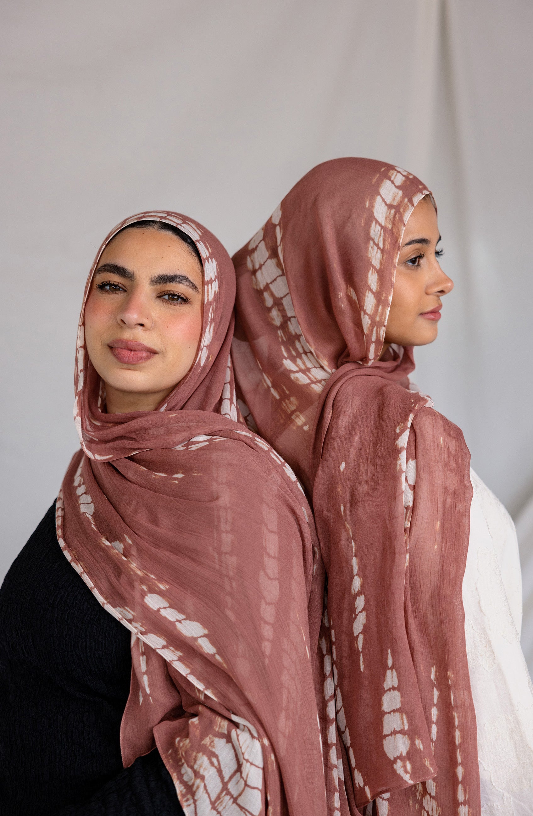 Two women wearing matching pink hijabs with a subtle white ripple pattern against a plain background