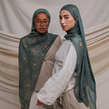 Two models wearing a teal woven hijab with crescents on it. the hijab is draped on the models around their head. the models are standing in front of a cream fabric backdrop.