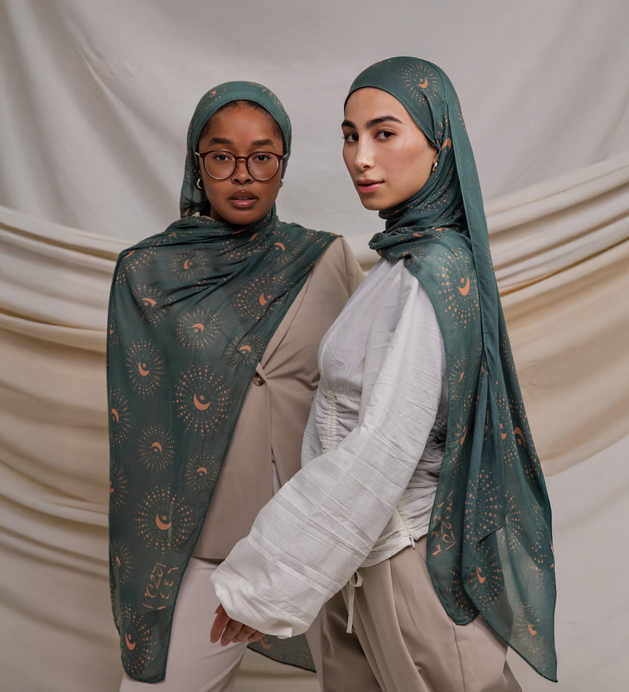 Two models wearing a teal woven hijab with crescents on it. the hijab is draped on the models around their head. the models are standing in front of a cream fabric backdrop.