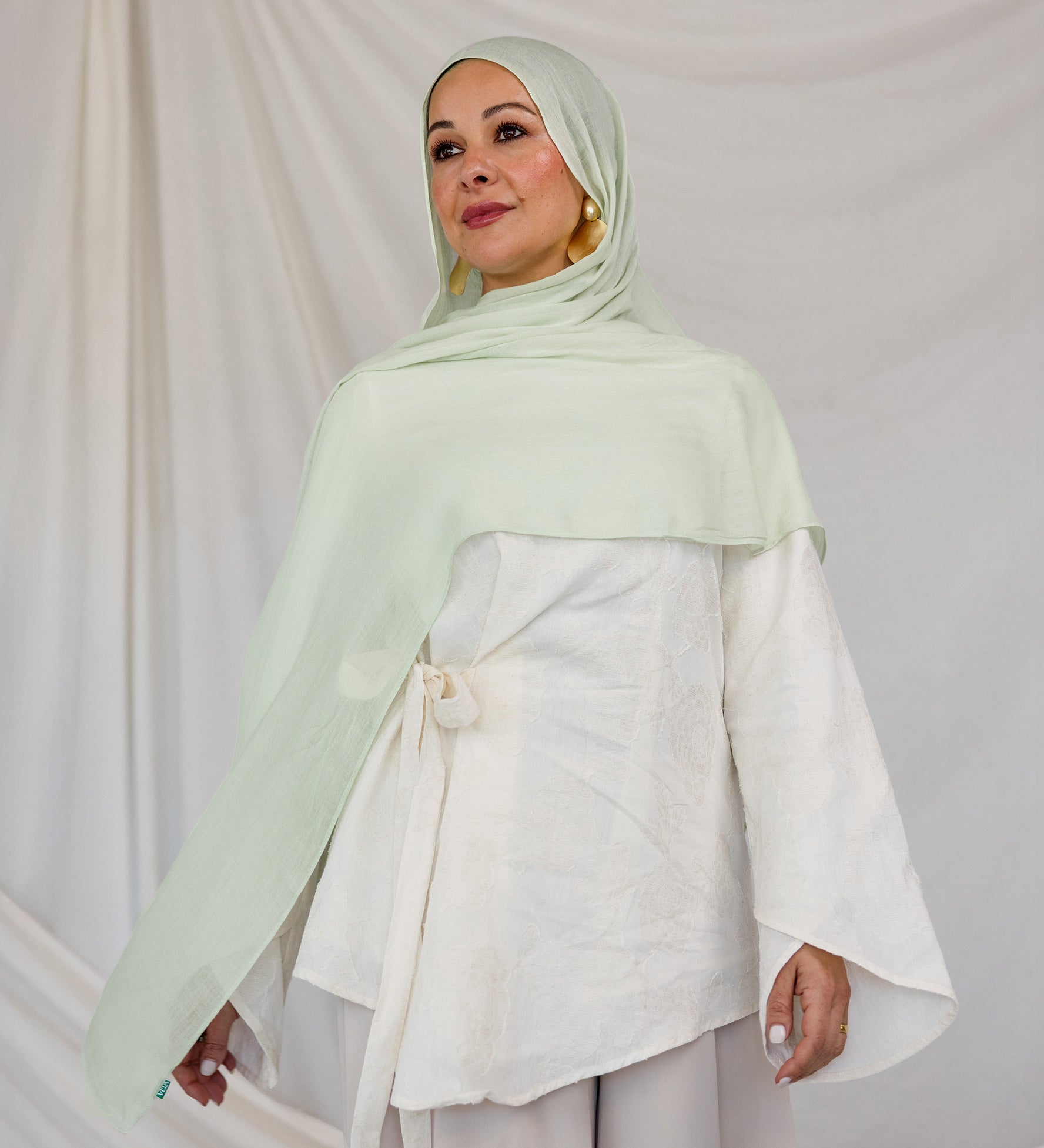 Model styled in a light pastel green hijab and white outfit against a plain background