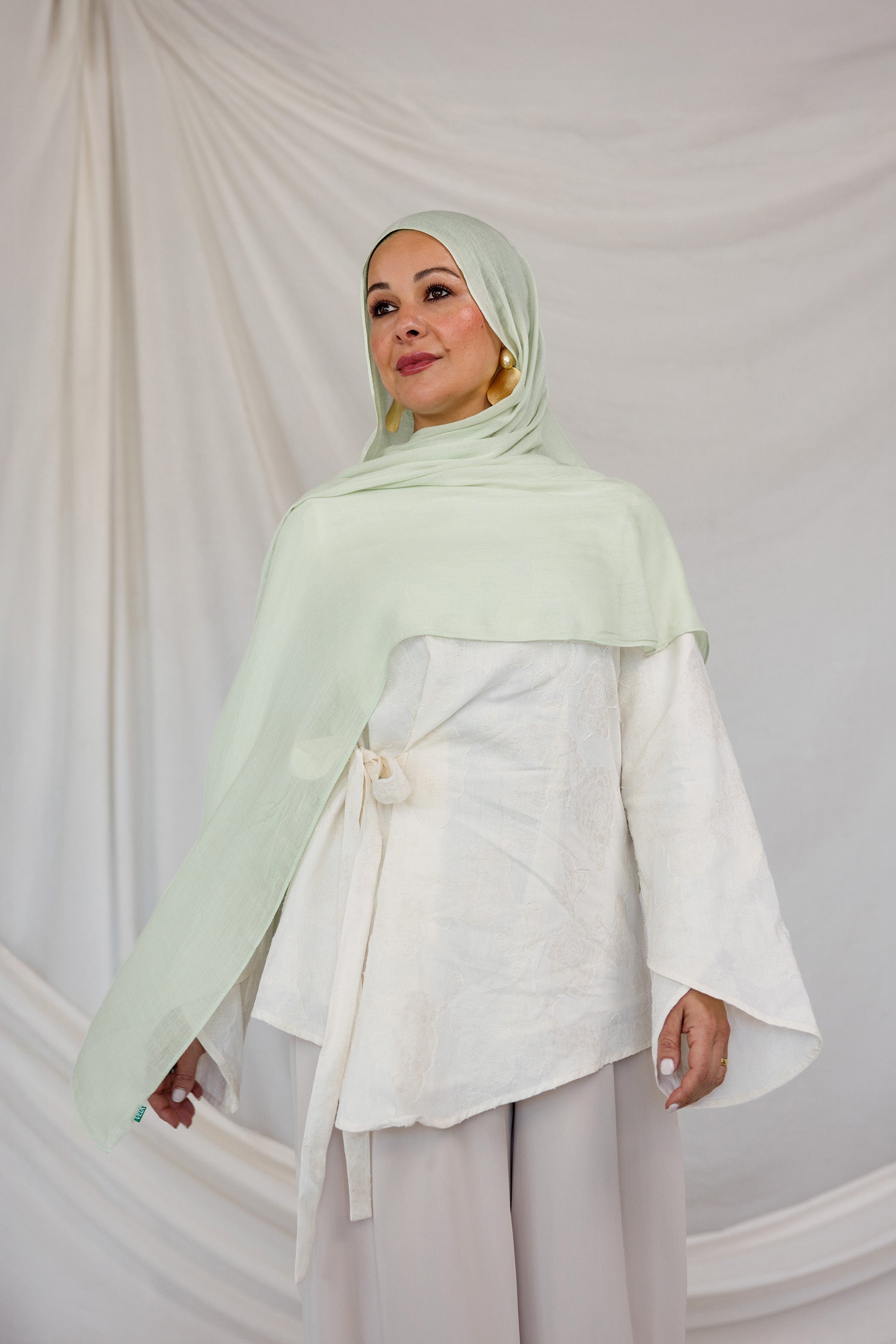Model styled in a light pastel green hijab and white outfit against a plain background