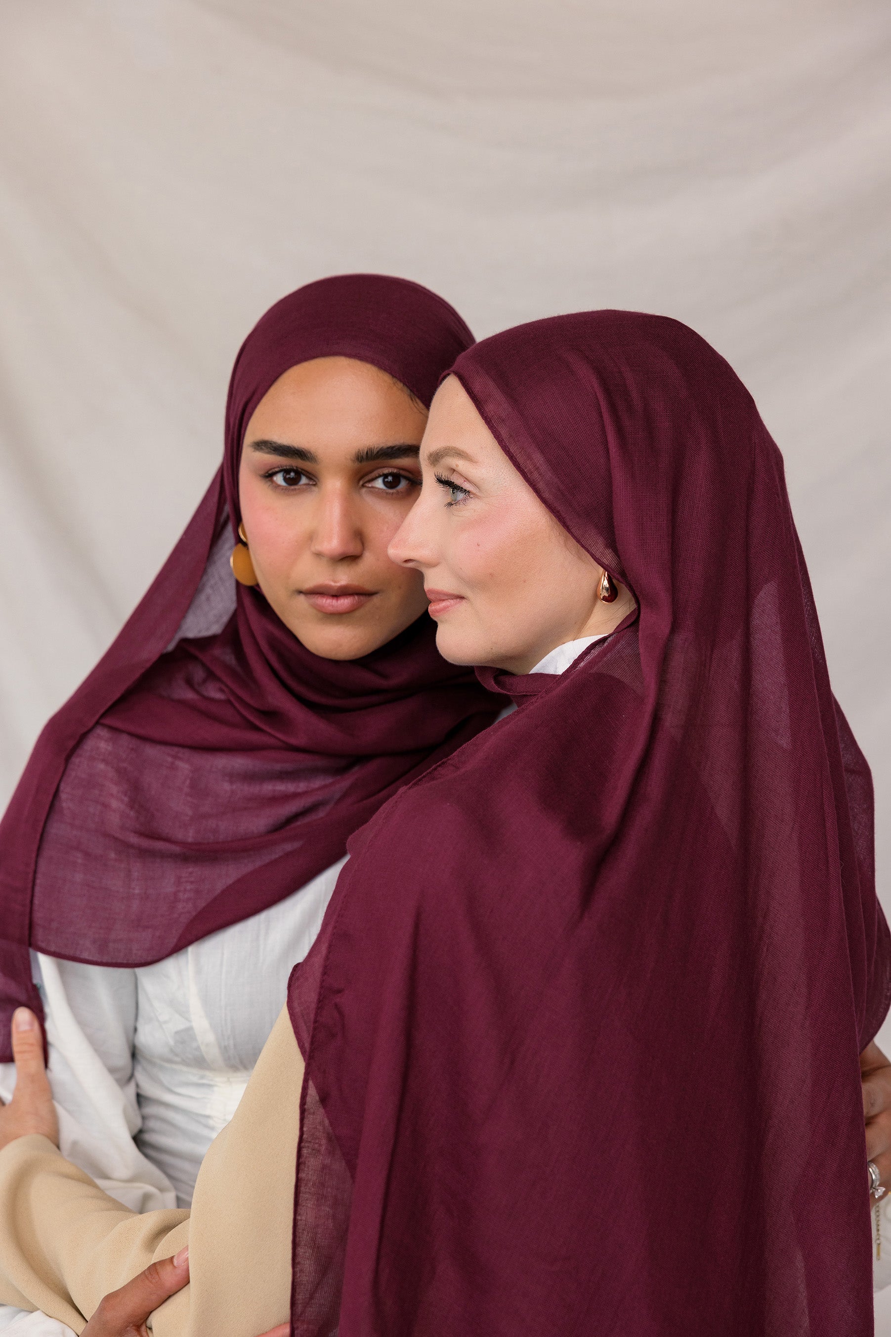 Two models standing side by side wearing the Maroon hijab, styled neatly to frame the face against a neutral studio backdrop.