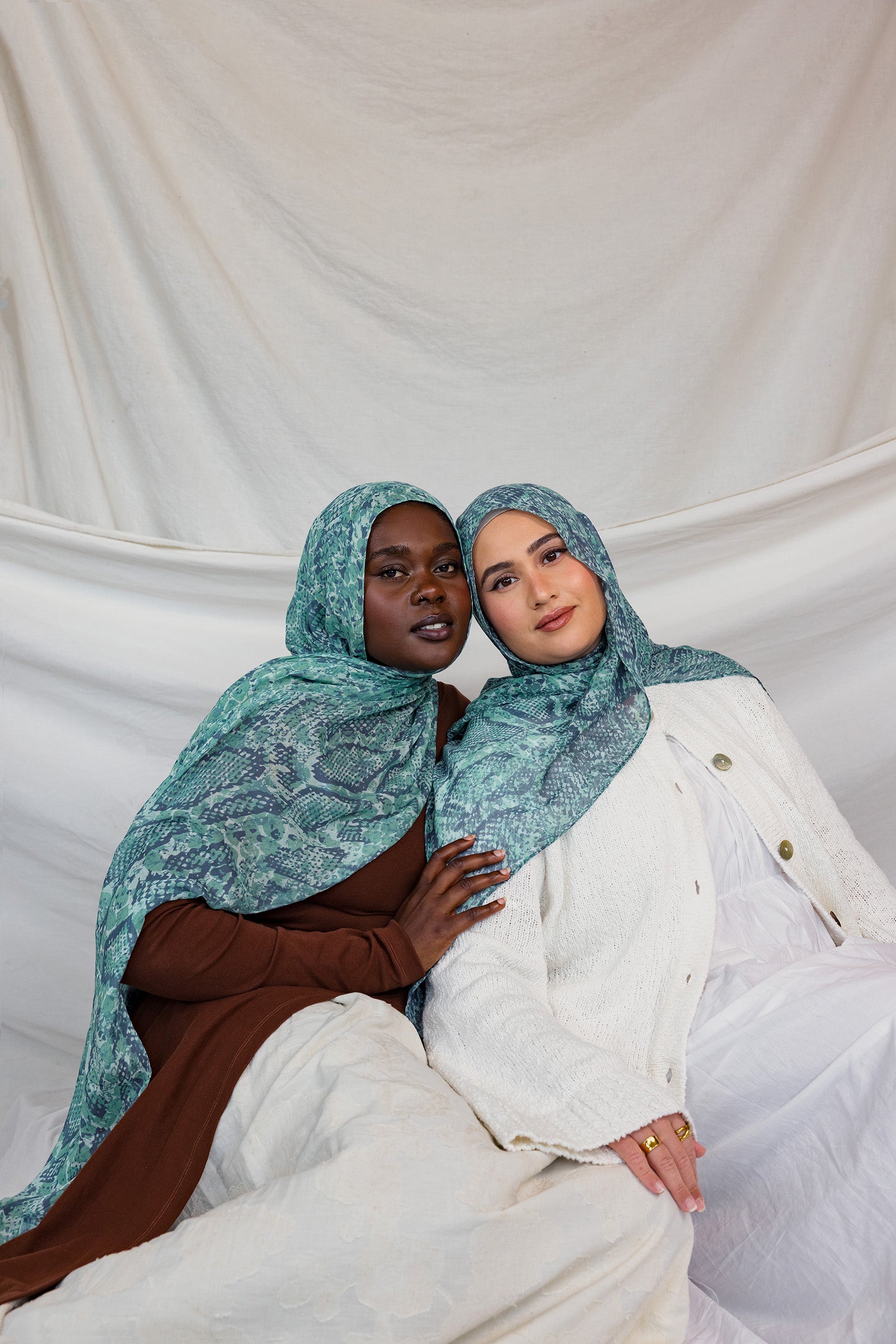 Two models shown wearing the Aqua Cobra scarf by VELA, featuring a snakeskin print in teal and blue.