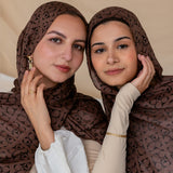 Two women wearing a brown hijab with Art Deco patterns, posing closely together with one holding her hijab away from her face.