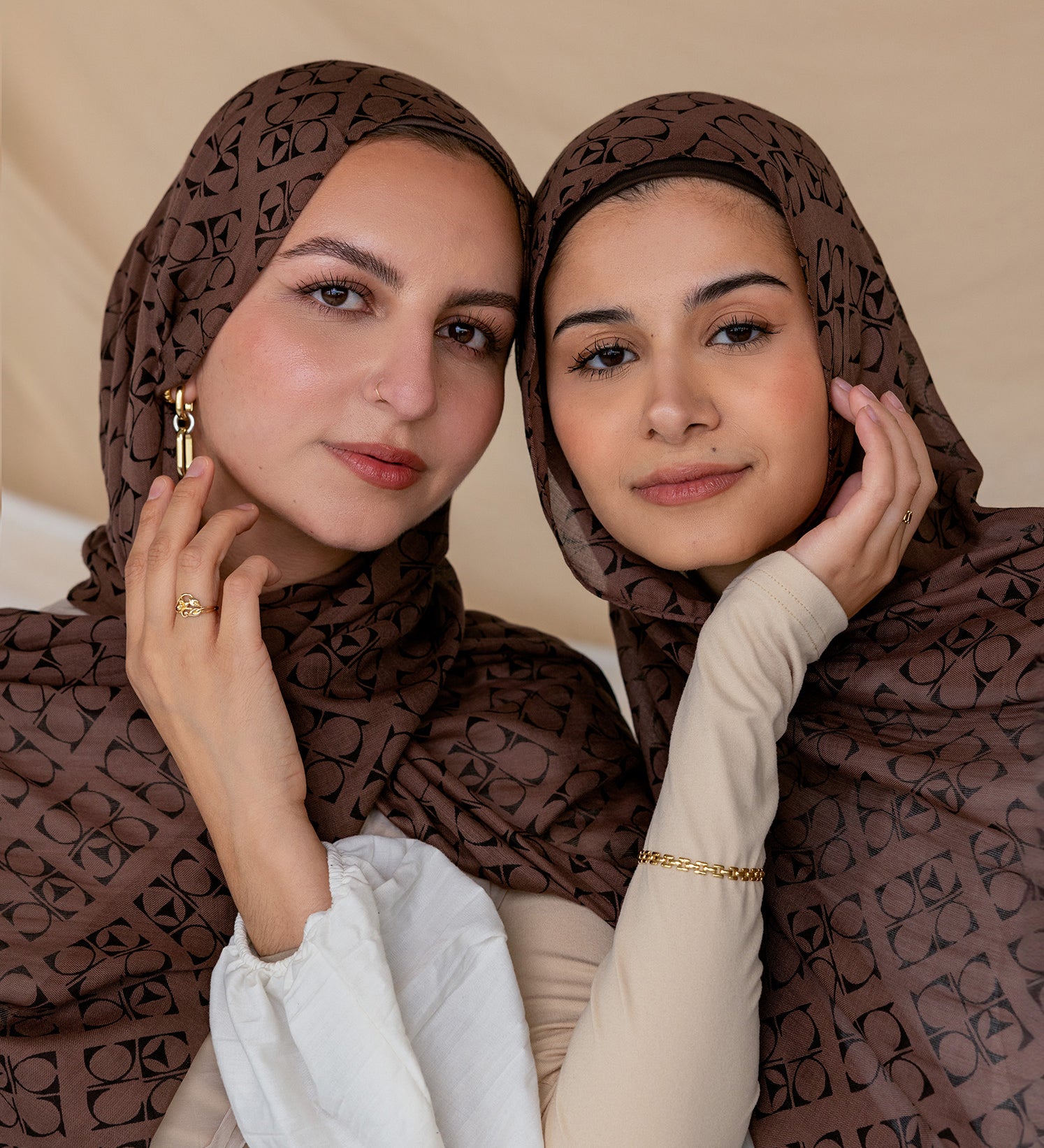 Two women wearing a brown hijab with Art Deco patterns, posing closely together with one holding her hijab away from her face.
