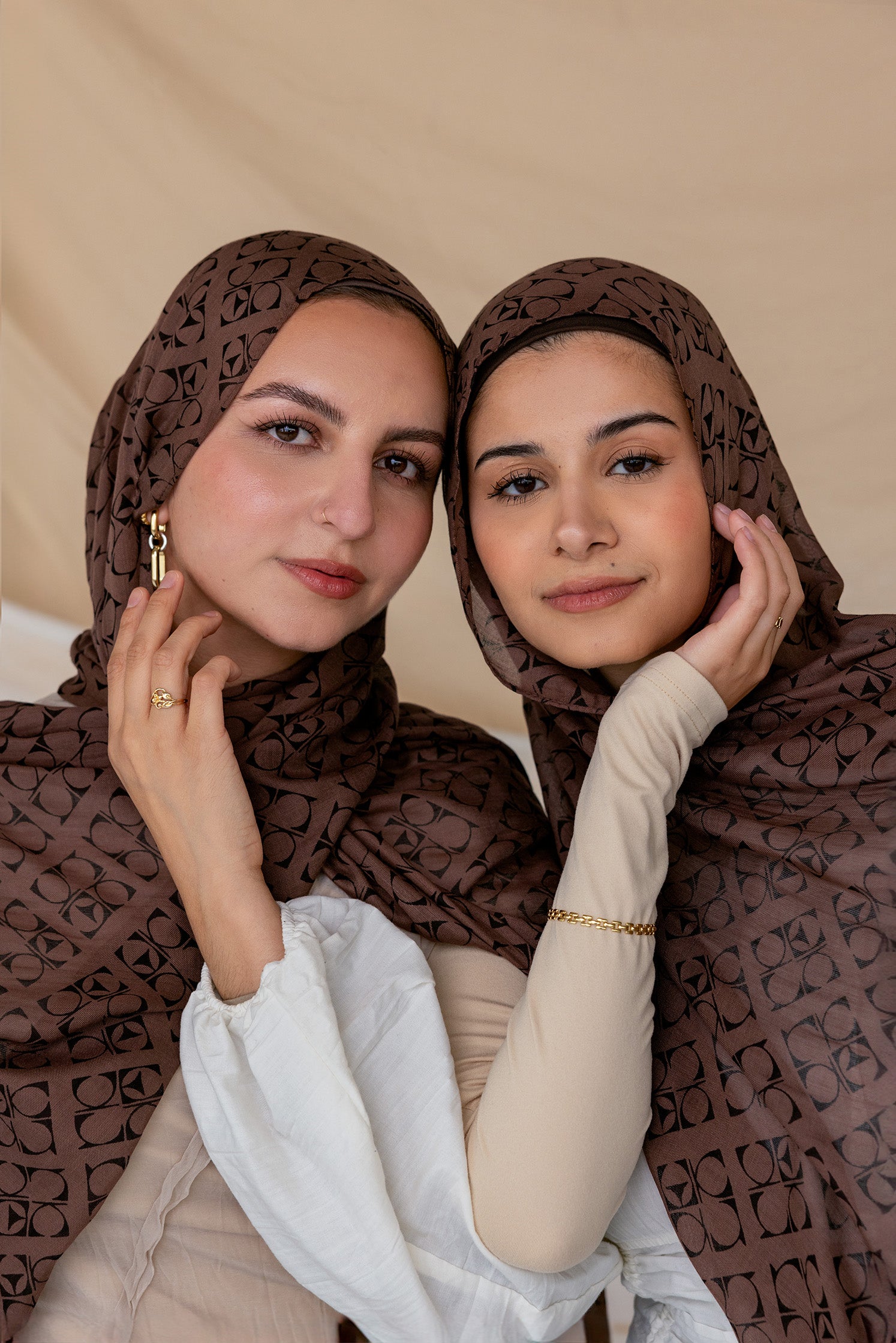 Two women wearing a brown hijab with Art Deco patterns, posing closely together with one holding her hijab away from her face.