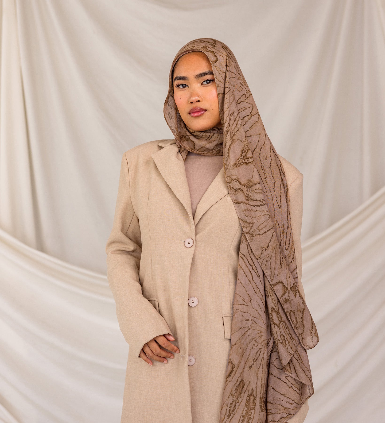 Woman wearing a beige coat and mocha patterned hijab against a plain background