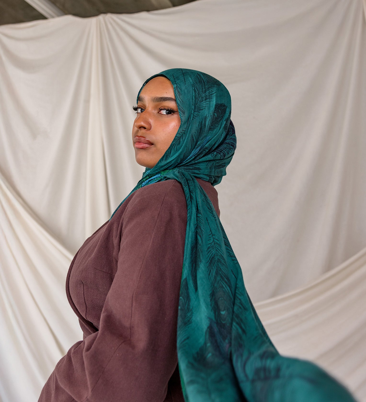 Model wearing a teal peacock inspired printed hijab and brown top against a white backdrop