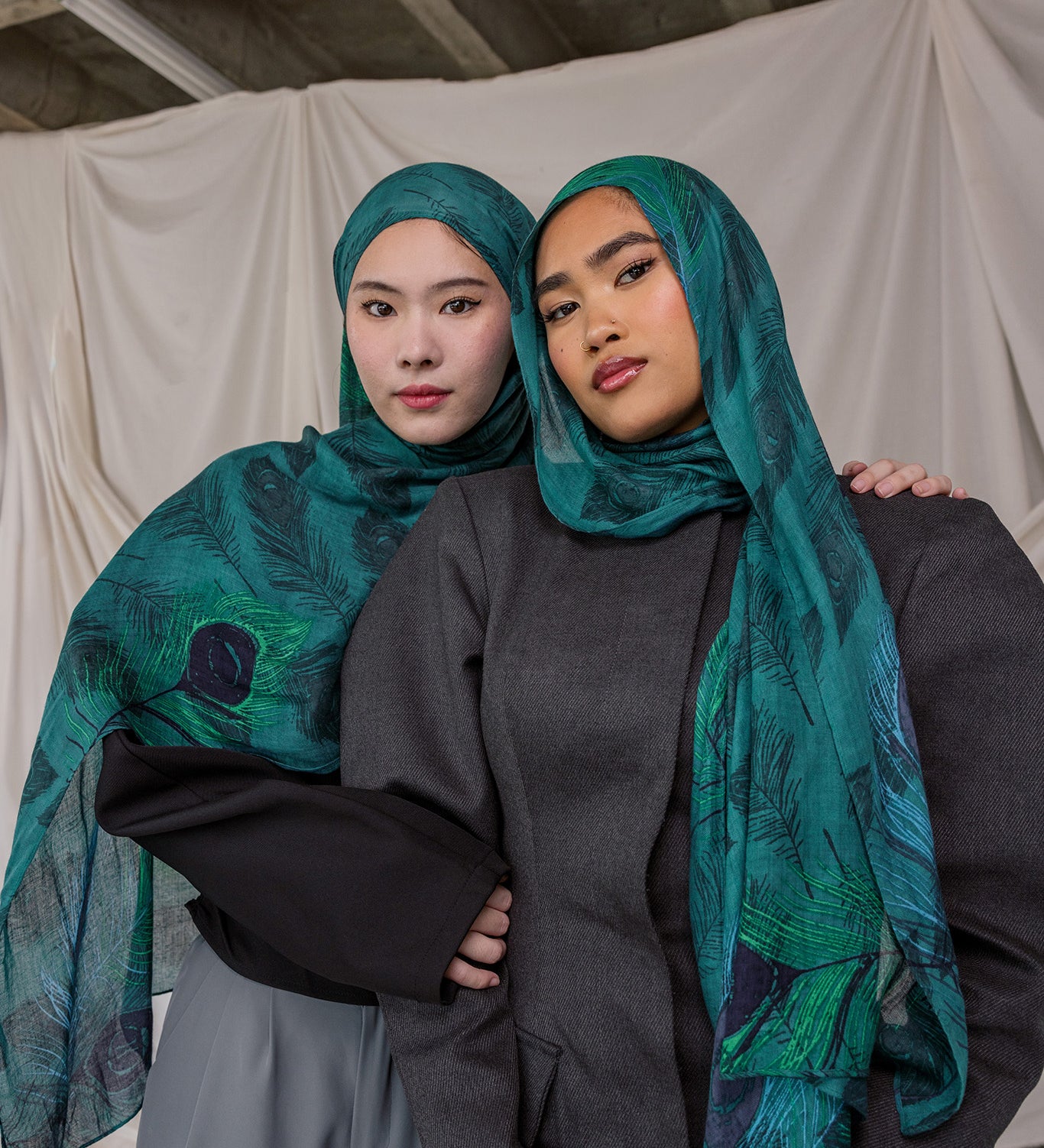 Two women wearing peacock feather inspired headscarves and dark clothing against a plain background