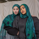 Two women wearing peacock feather inspired headscarves and dark clothing against a plain background