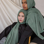 Two women wearing Pine Green hijabs sitting on a white draped background