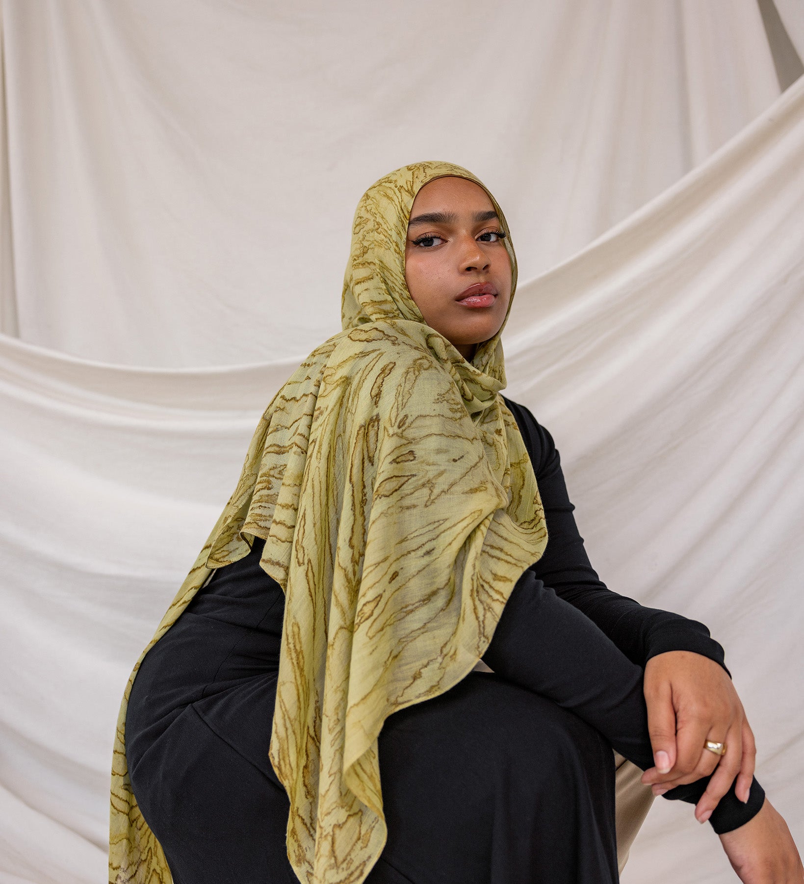 Model wearing a olive printed dune hijab and black outfit against a white draped background