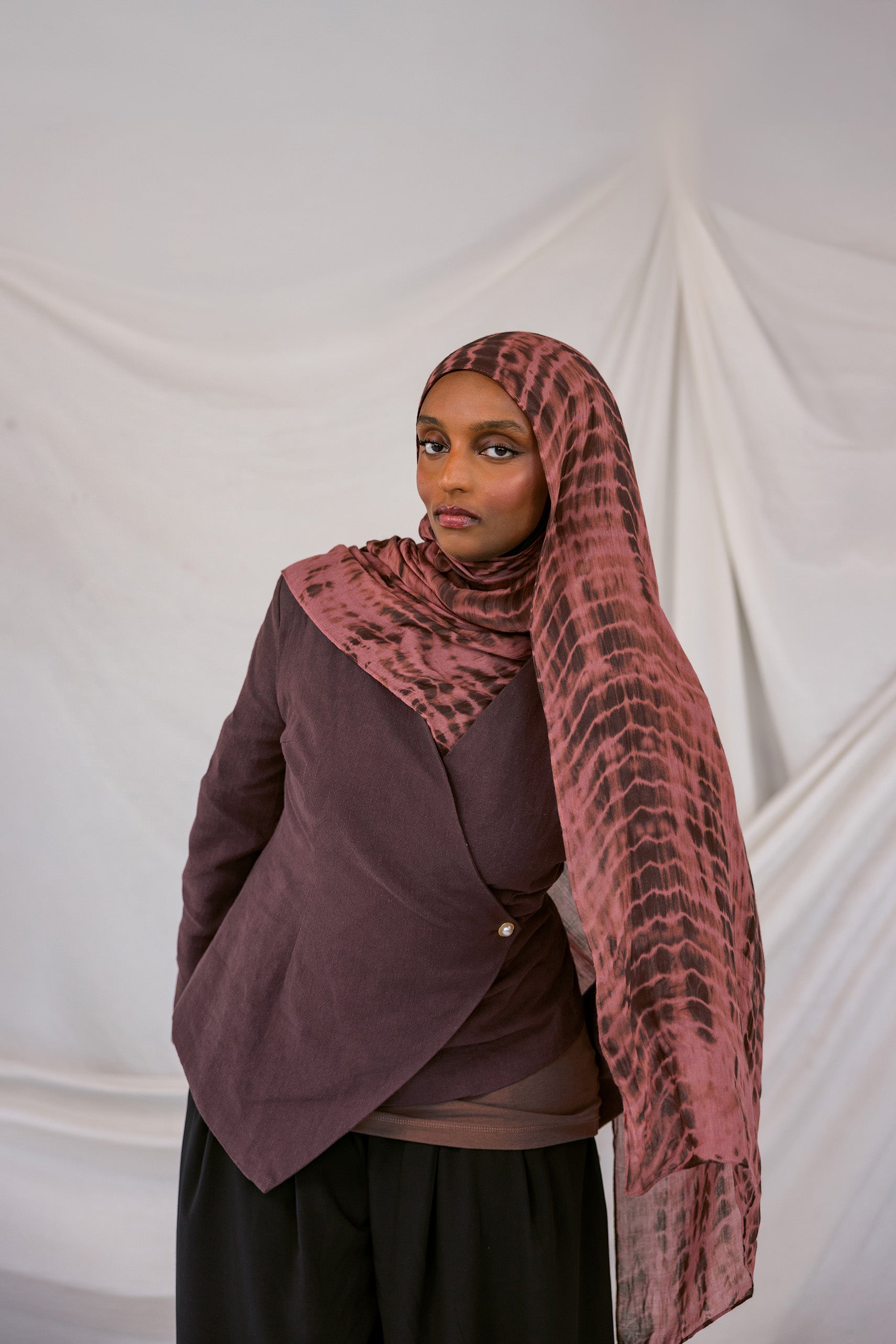 Model wearing a red clay ripple patterned hijab and dark clothing against a plain background
